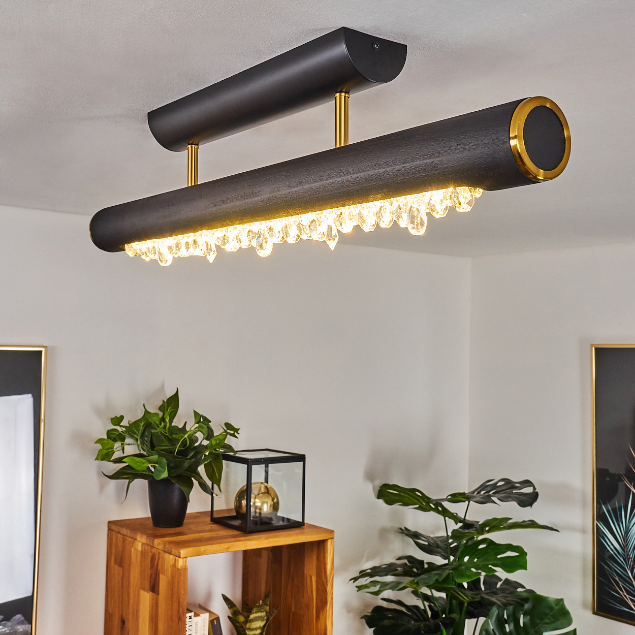 Moderne schwarze Deckenleuchte mit Kristallakzenten. Darunter ein Holzregal mit Pflanzen und Dekorationsartikeln. Helles Interieur.