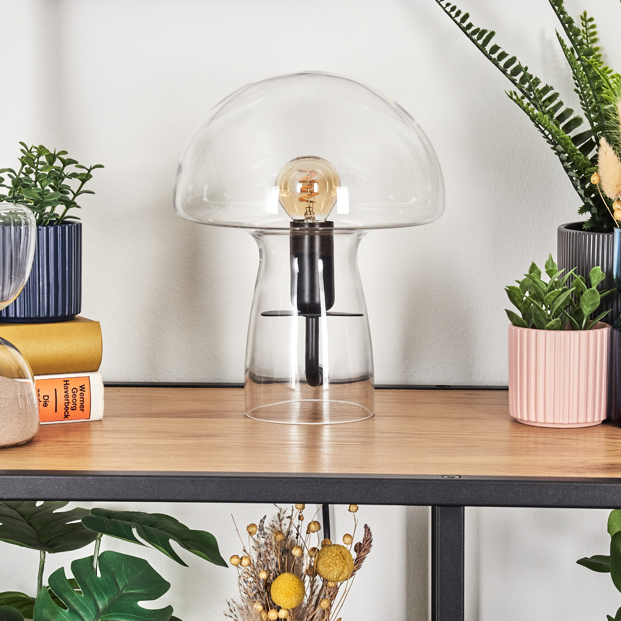 Eine gläserne Pilzlampe steht auf einem Holzregal, flankiert von Topfpflanzen und Büchern. Getrocknete Blumen und Grün sind darunter zu sehen.