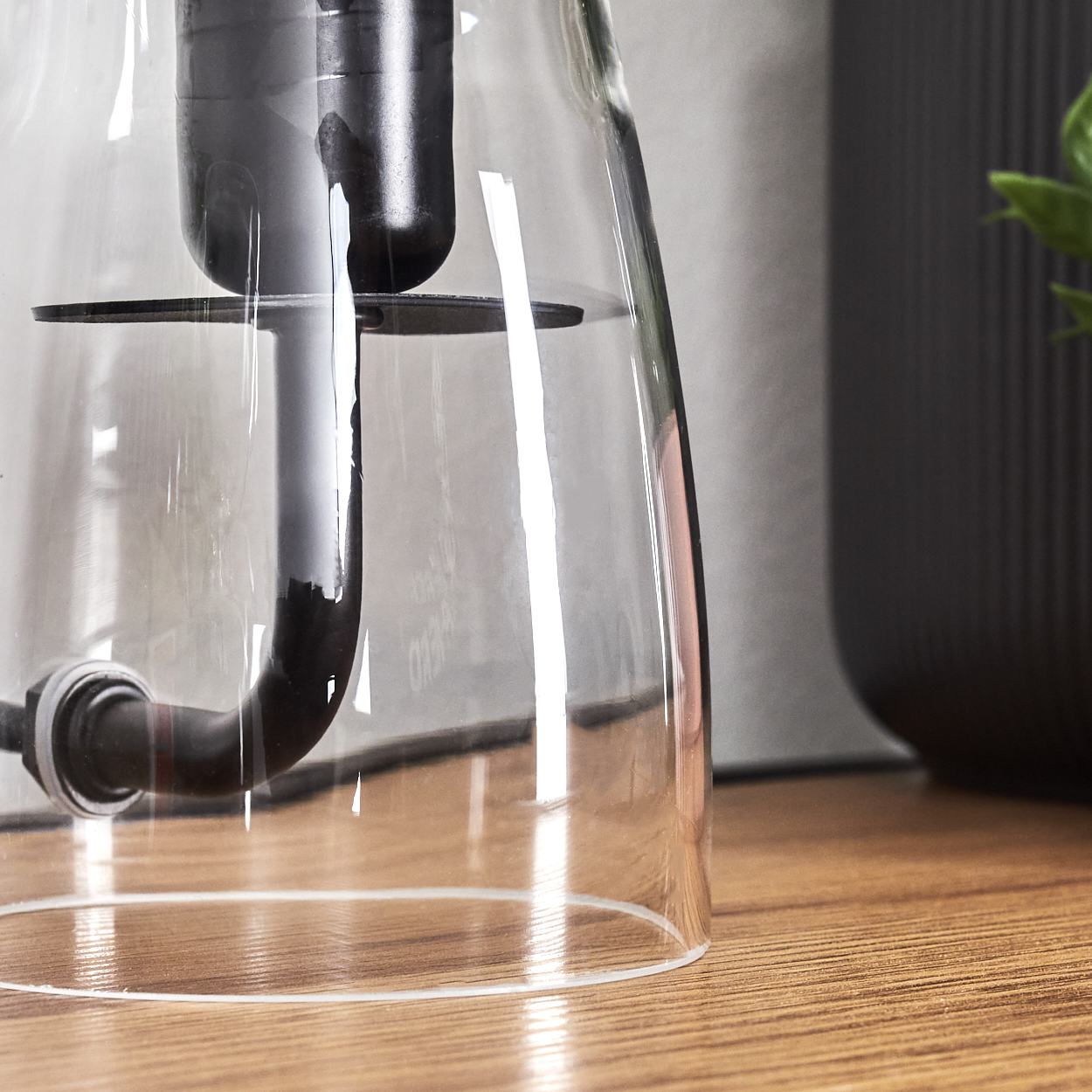 Nahaufnahme einer Glaskaraffe mit schwarzen Komponenten. Sie steht auf einer Holzoberfläche, im Hintergrund eine dunkle Vase und Pflanze.