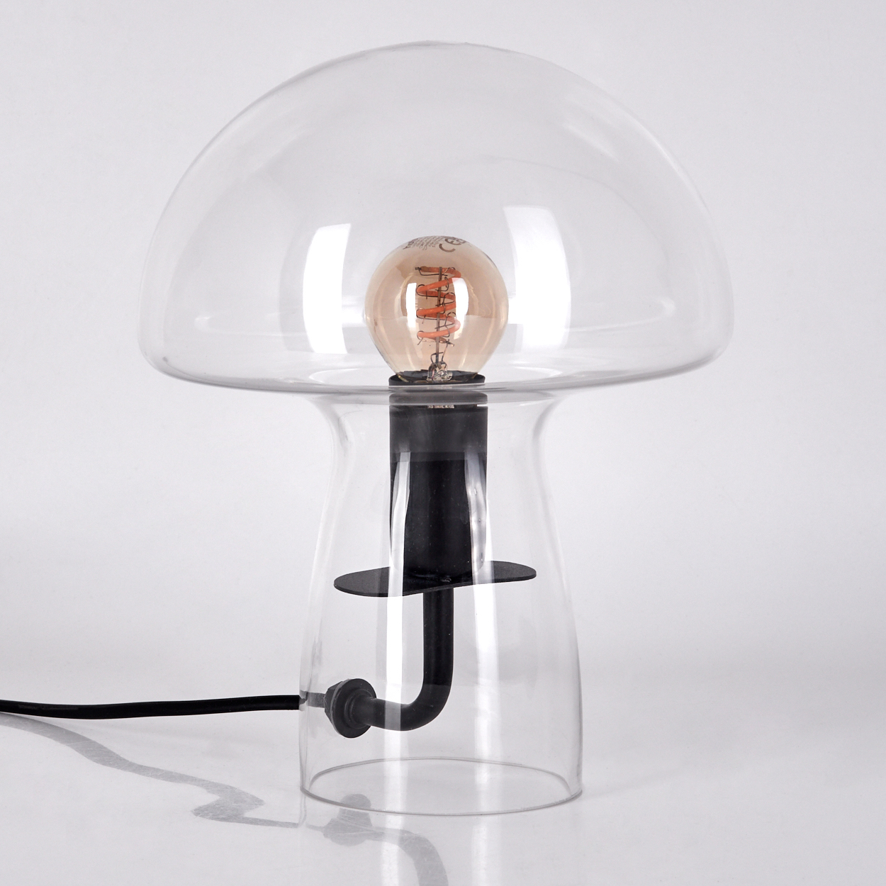 Eine pilzförmige Tischlampe mit klarem Glasschirm und -fuß, schwarzem Stiel und sichtbarer Glühbirne.
