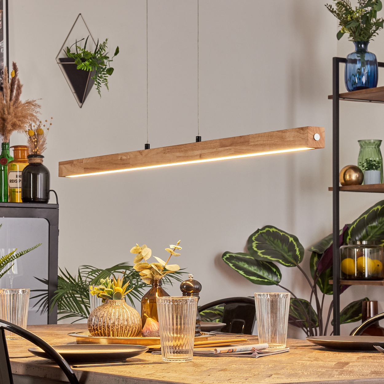 Holzleuchte über einem Tisch mit Gläsern und Tellern. Pflanzen und Dekorationen sind im Hintergrund sichtbar.