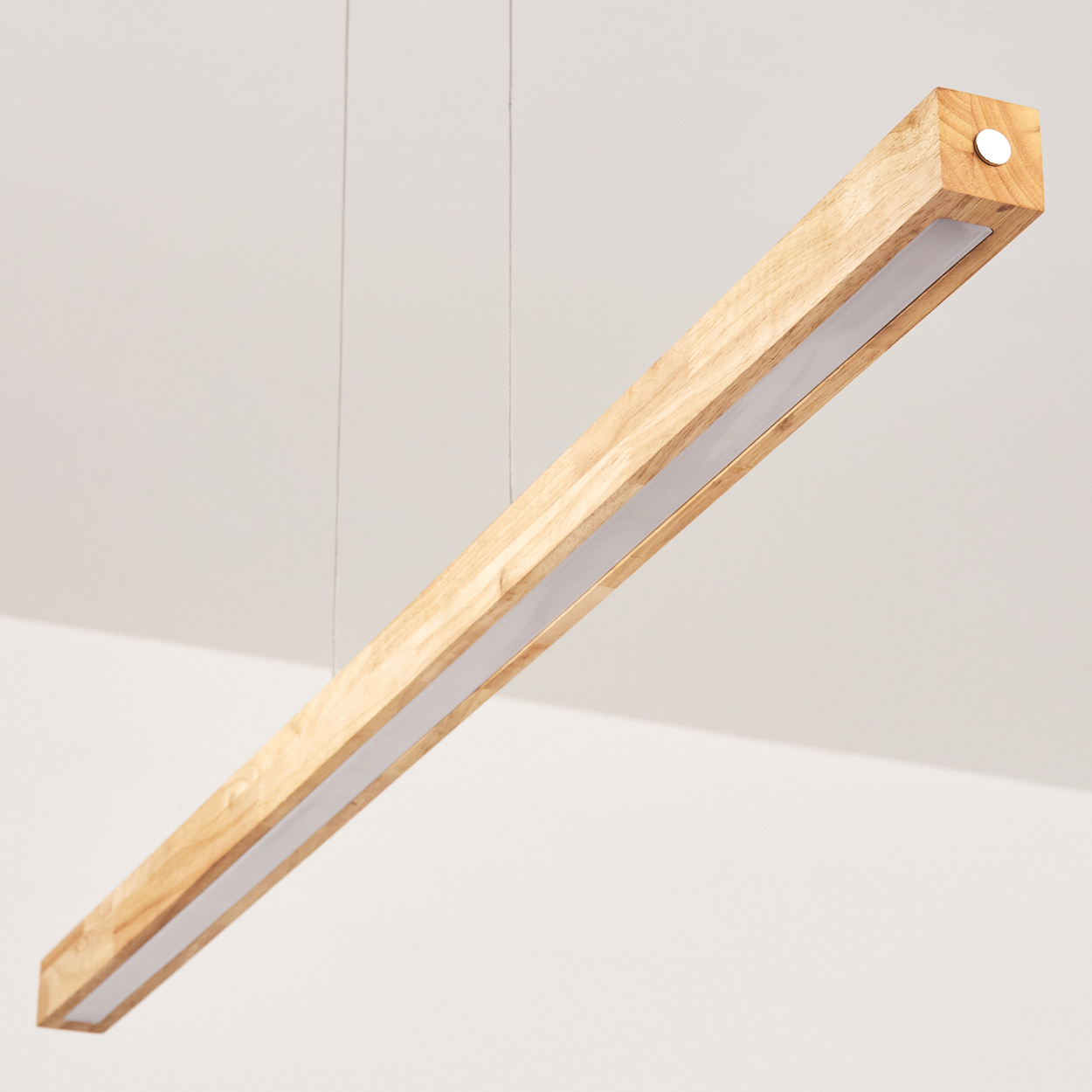 Eine rechteckige Holzleuchte hängt an dünnen Drähten. Sie hat eine weiße Lichtfläche und hängt an einer weißen Decke.