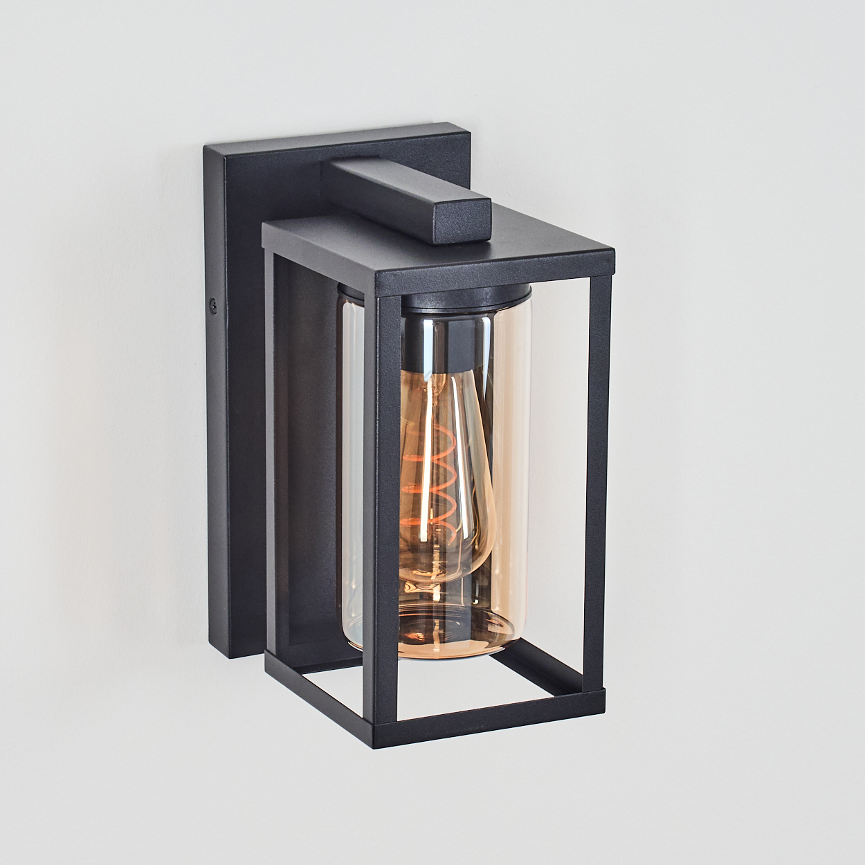 Eine schwarze Wandleuchte mit einem durchsichtigen Glaszylinder in einem rechteckigen Rahmen, der eine warmtonige Glühbirne beherbergt.