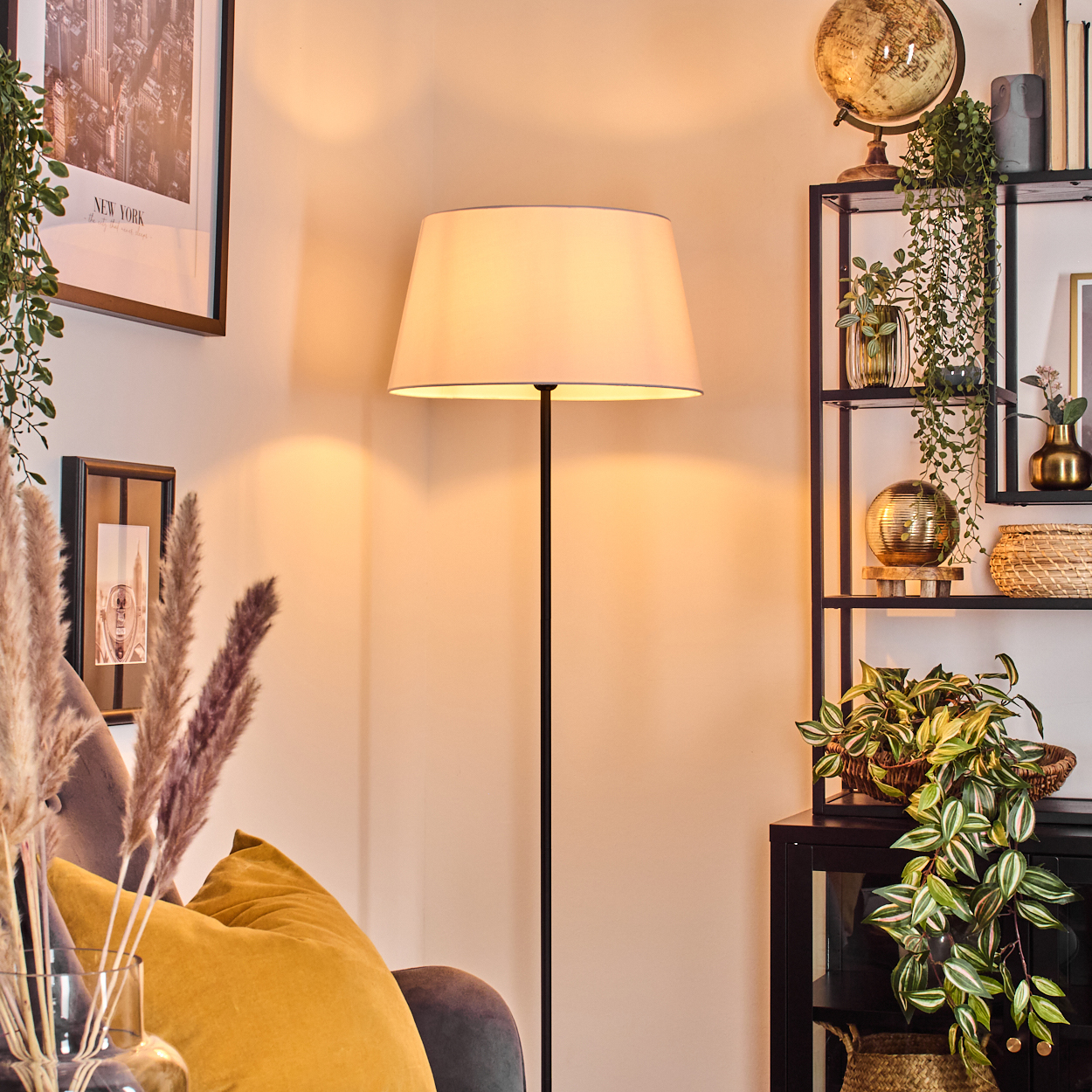 Ein Wohnzimmer mit Stehlampe, Regal mit Deko und Pflanzen und einem gelben Kissen auf einer Couch.