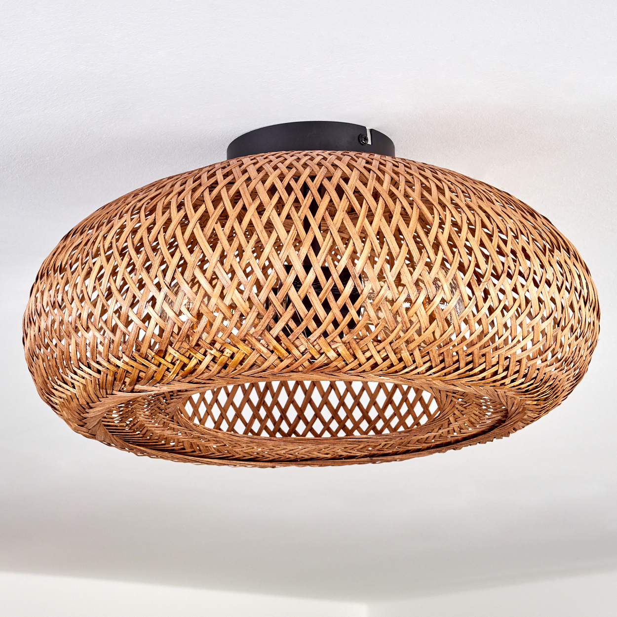 Eine gewebte Pendelleuchte aus Weide, oval geformt, hängt von einer weißen Decke. Die Lampe hat einen dunklen Sockel. Das Geflecht ist hellbraun.