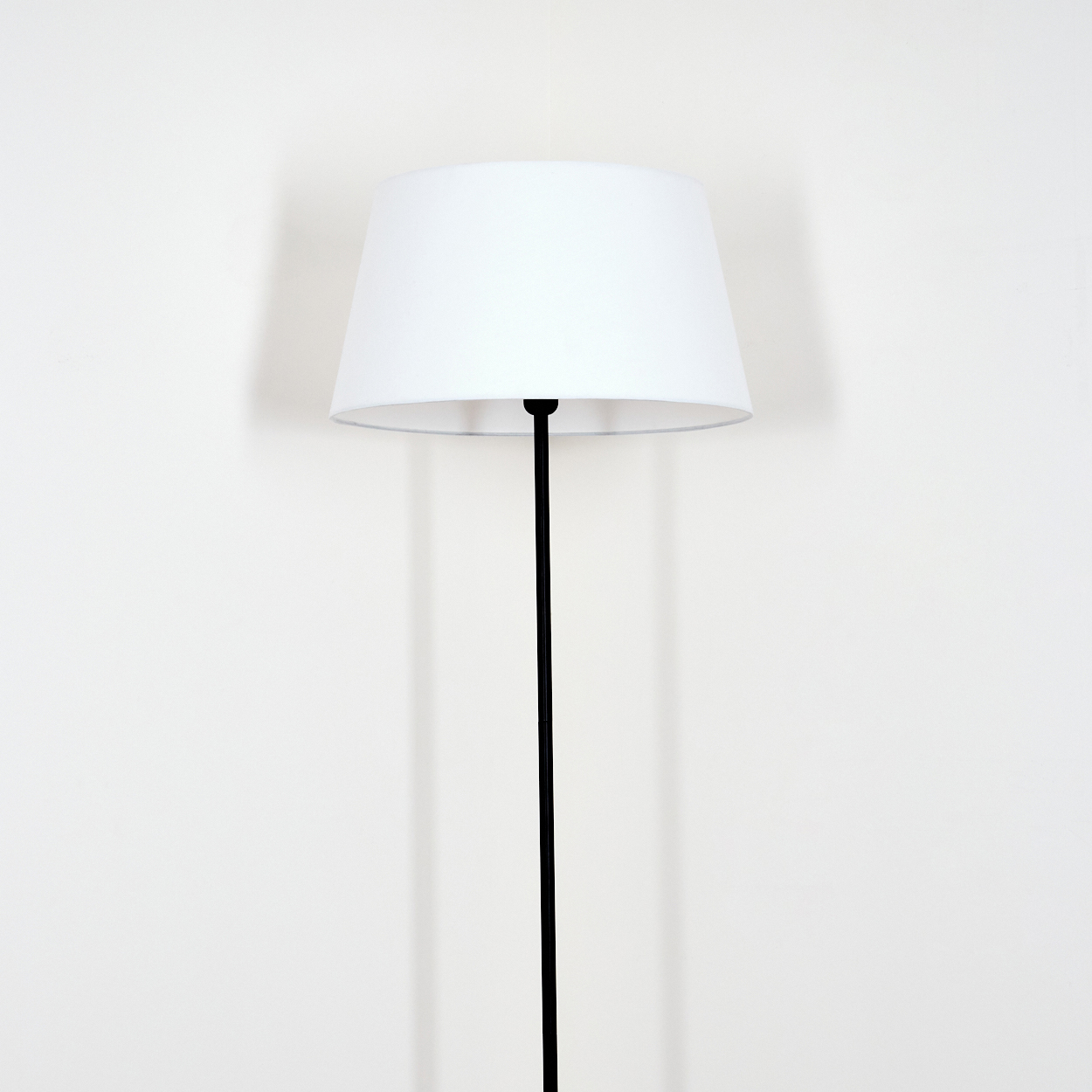 Ein weißer Lampenschirm sitzt auf einem schwarzen Pfahl. Er wirft einen Schatten auf die weiße Wand dahinter. Die Lampe ist mittig im Bild.