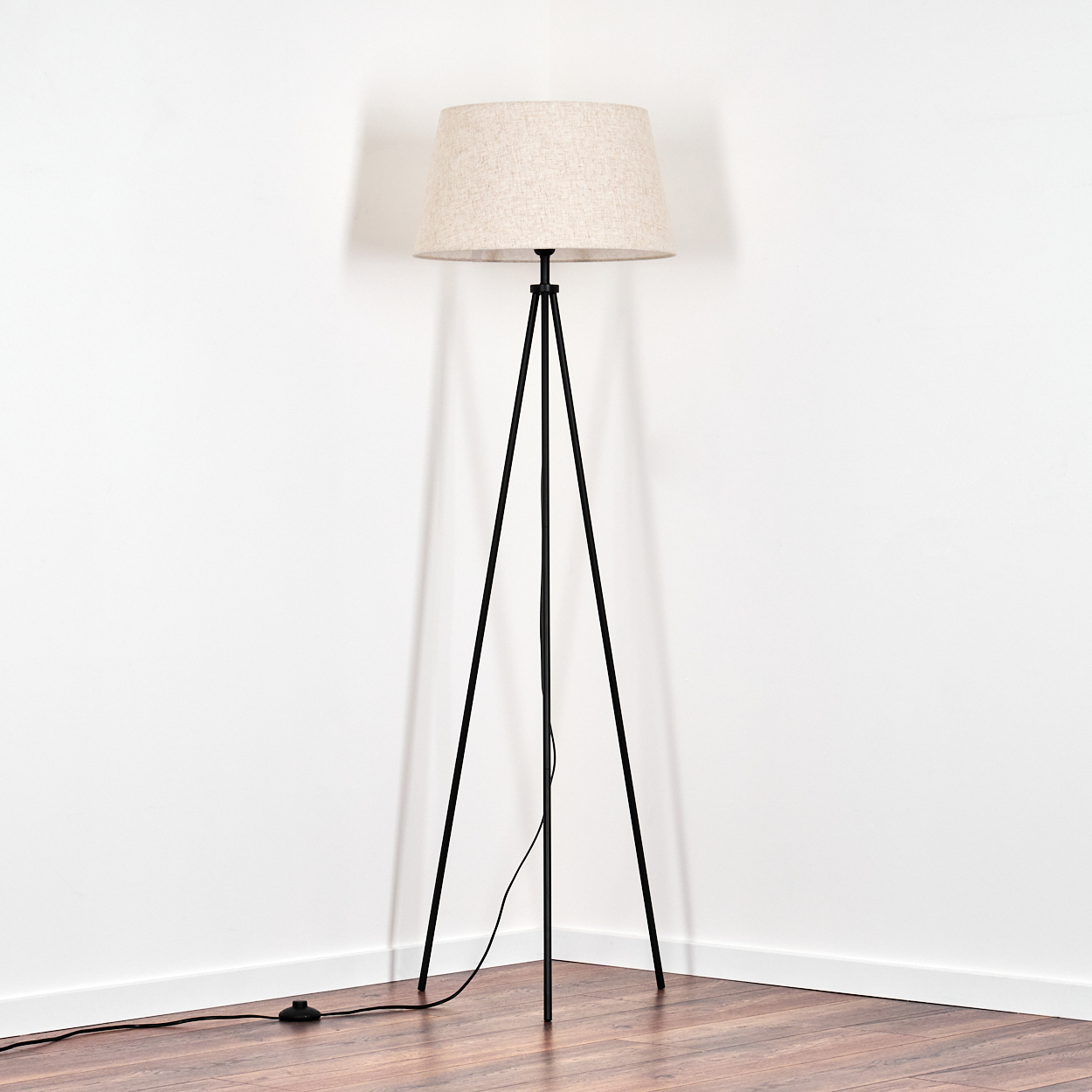 Eine schwarze Stehleuchte mit beigem Lampenschirm steht in einer Ecke vor weißen Wänden und einem Holzboden.