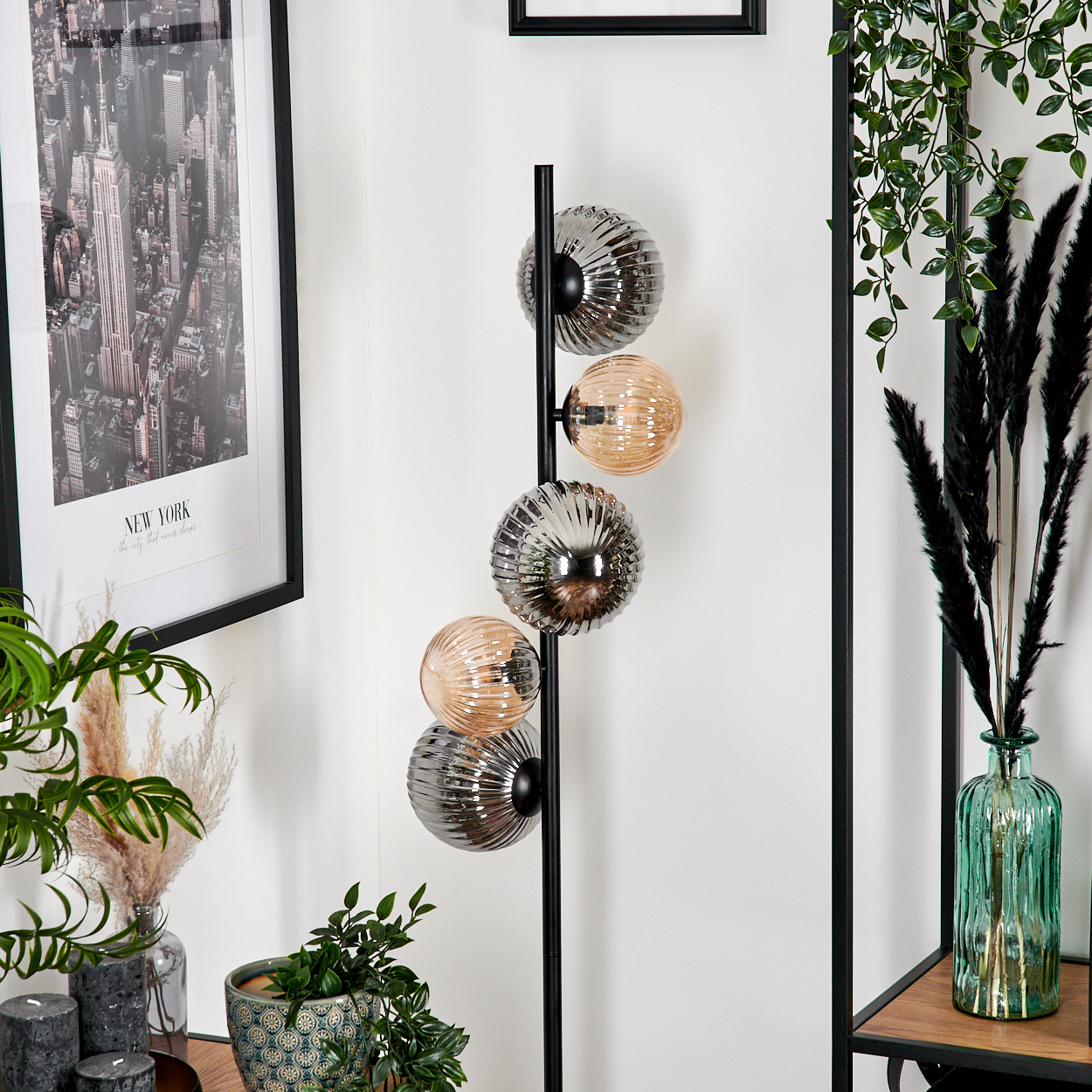 Eine Stehlampe mit Kugelschirmen, ein gerahmter New York-Druck und Pflanzen in Vasen und Töpfen vor einer weißen Wand.