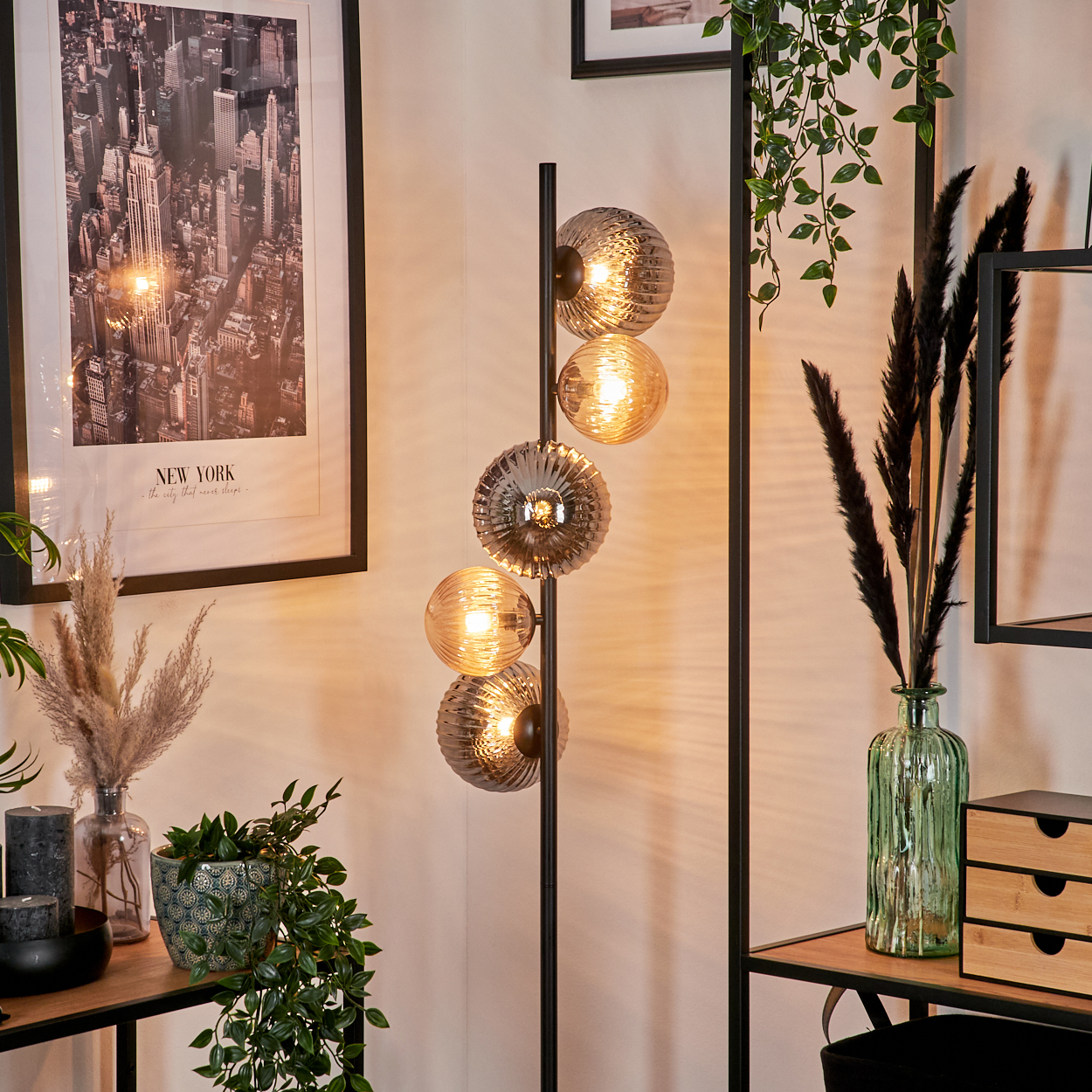 Zimmerecke mit Stehlampe mit runden Glasschirmen. Ein New York-Poster, Pflanzen und ein Holzregal mit Dekorationen sind sichtbar.