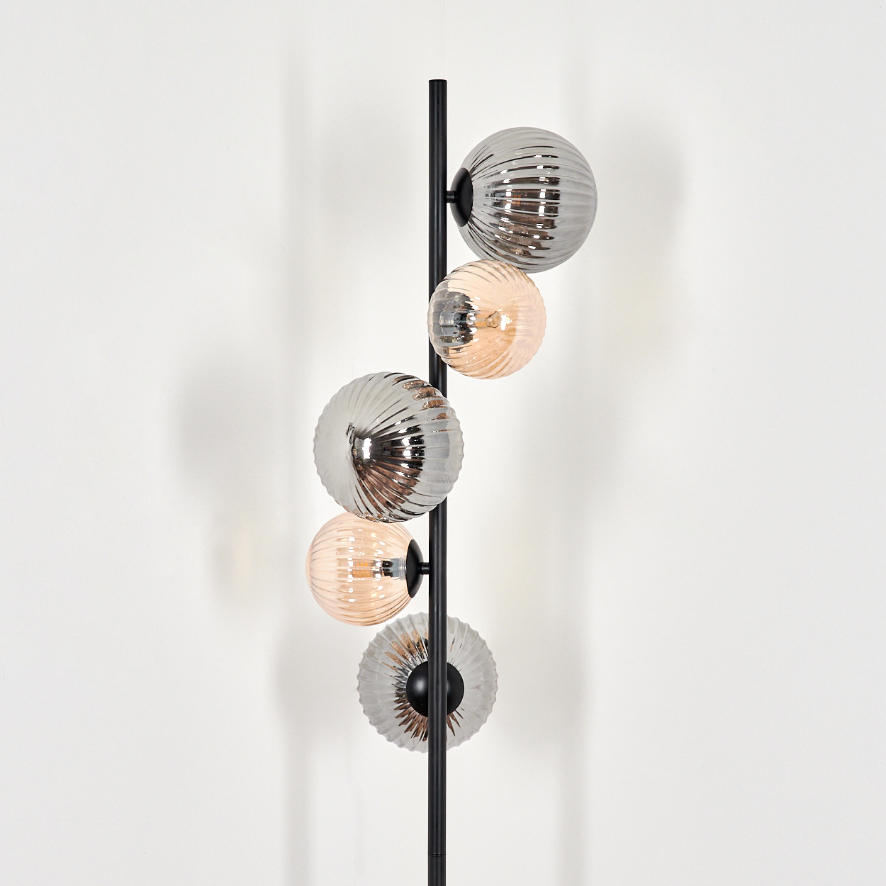 Eine schwarze Stehlampe mit fünf kugelförmigen Lichtern. Die Lichter wechseln sich zwischen Silber und Pfirsich ab, vor einer weißen Wand.
