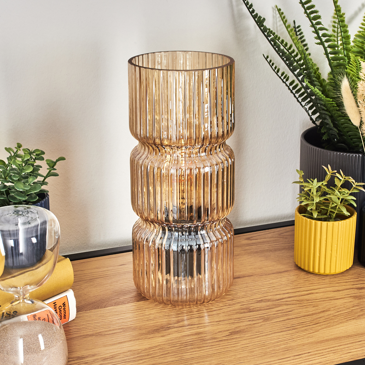 Bernsteinfarbene Glasvase auf einem Holzregal, Pflanzen in Töpfen, eine Sanduhr und Bücher. Die Vase hat ein geripptes Design.