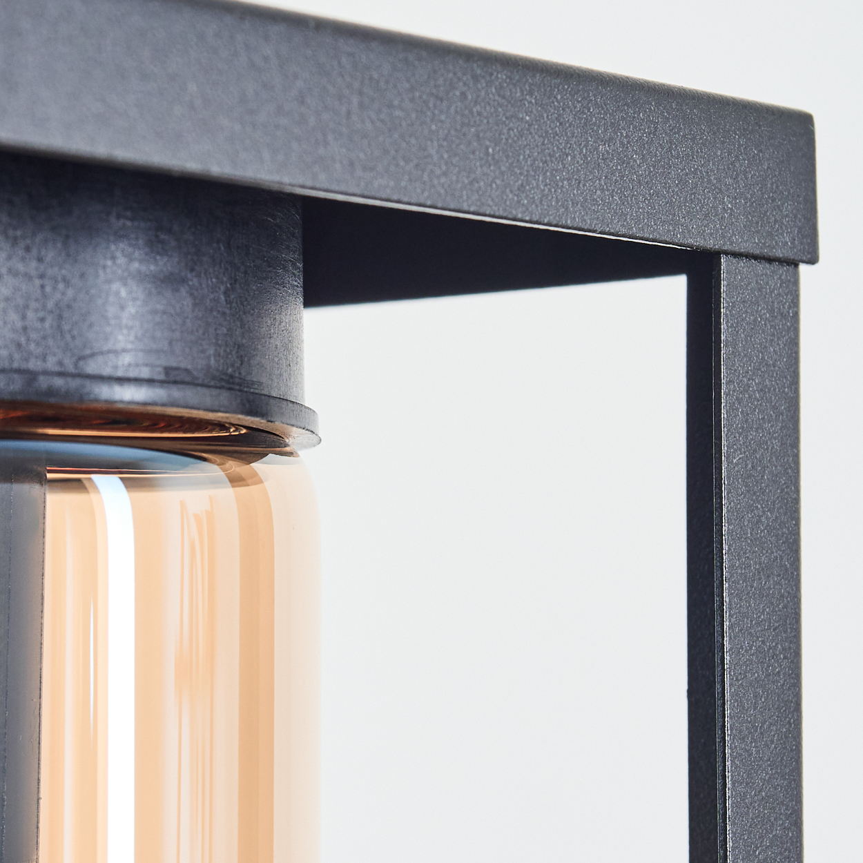 Nahaufnahme eines schwarzen Metallrahmens mit einem zylindrischen Glasobjekt. Das Glas ist bernsteinfarben, vor weißem Hintergrund.