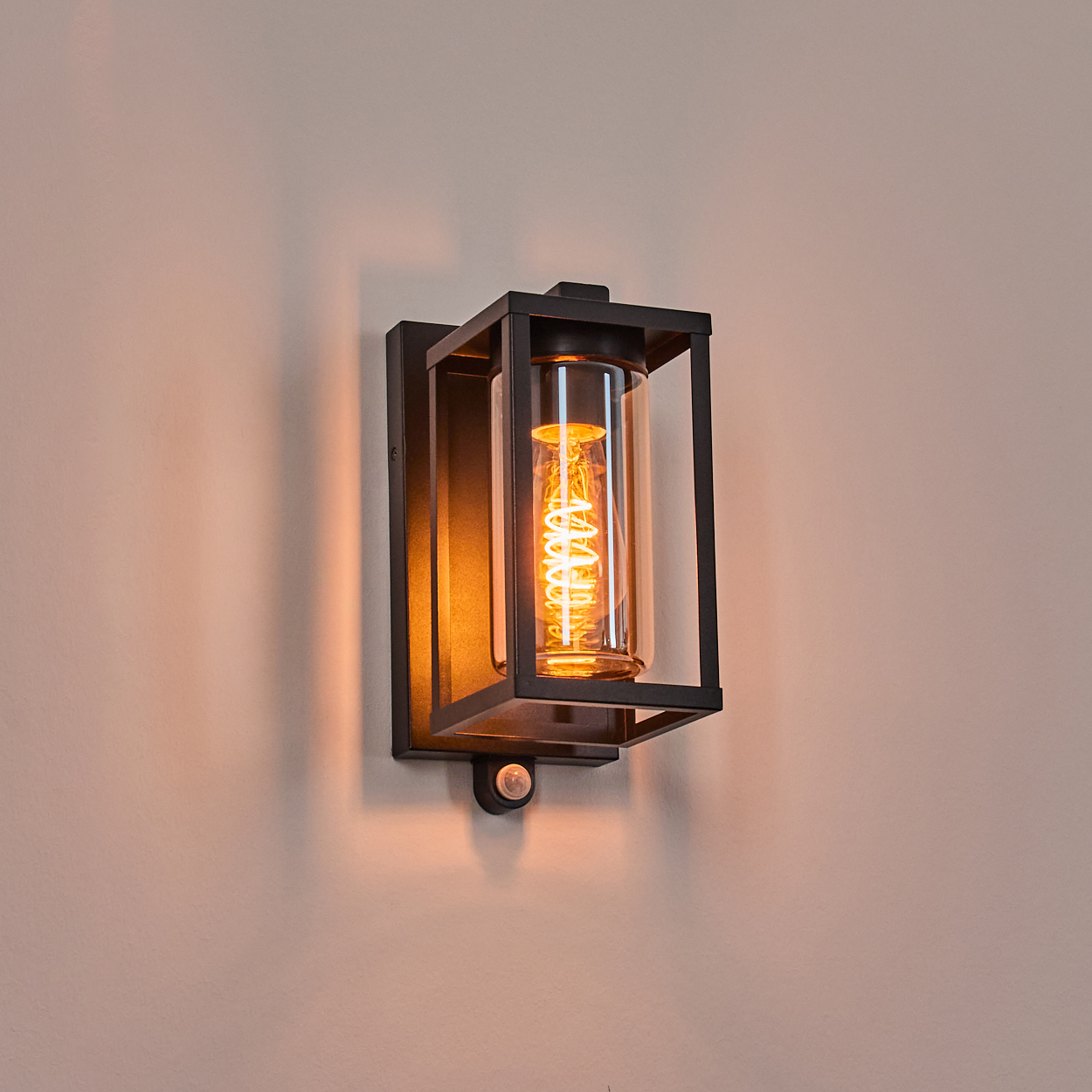 Eine moderne schwarze Wandlampe mit einem Glaszylinder, der eine Wendelglühbirne beherbergt, an einer hellrosa Wand montiert.