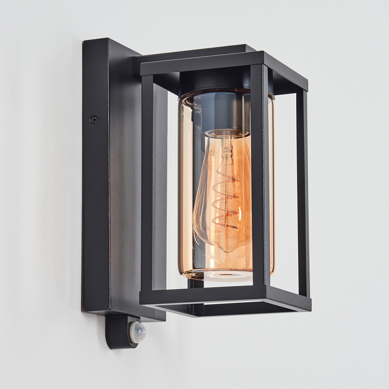 Schwarze Wandleuchte mit rechteckigem Rahmen und klarem Glaszylinder. Innen eine orangefarbene Glühbirne. Weißer Wandhintergrund.