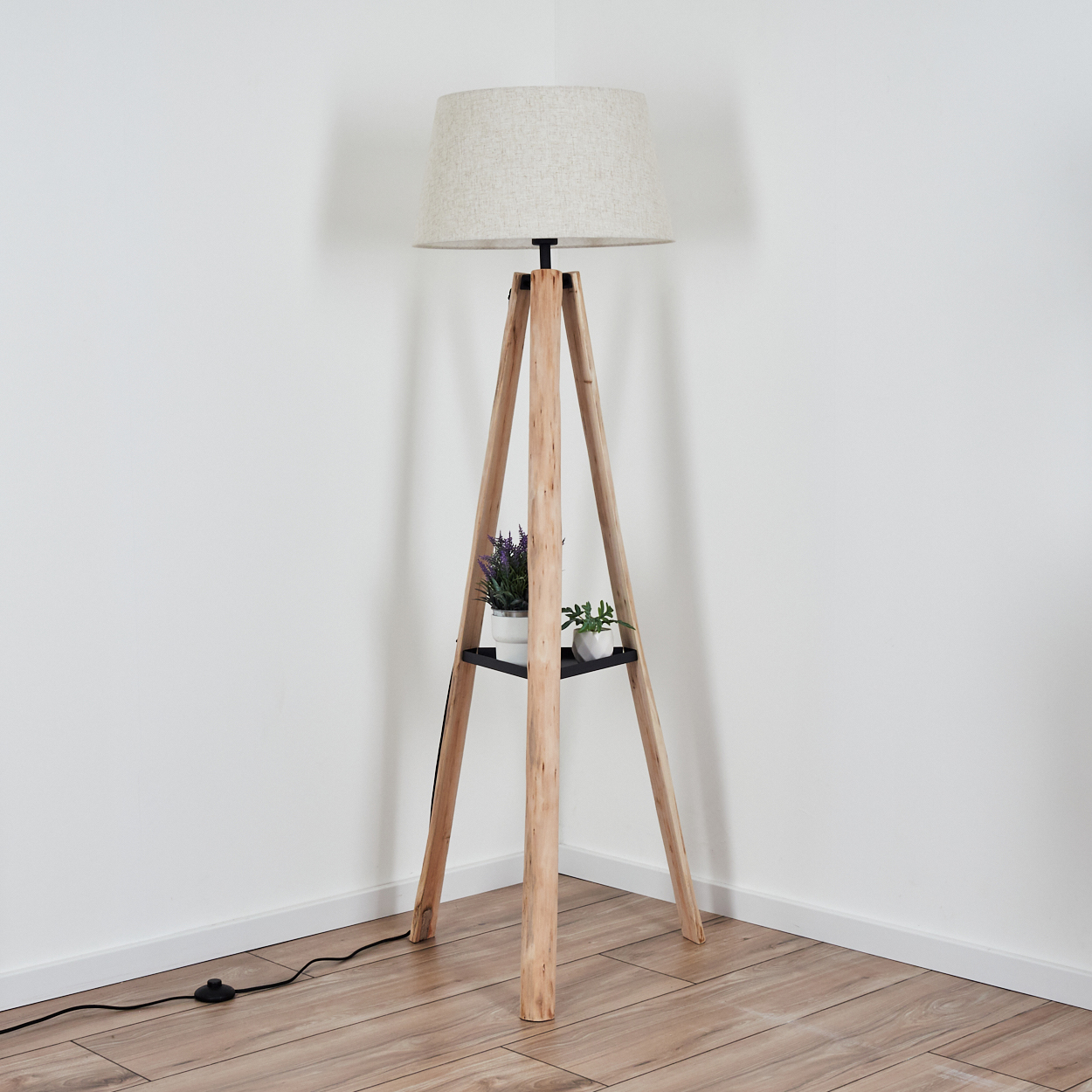 Eine hölzerne Stehlampe mit beigem Lampenschirm steht in einer Ecke. Sie hat ein Regal mit Topfpflanzen. Der Boden ist aus Holz.