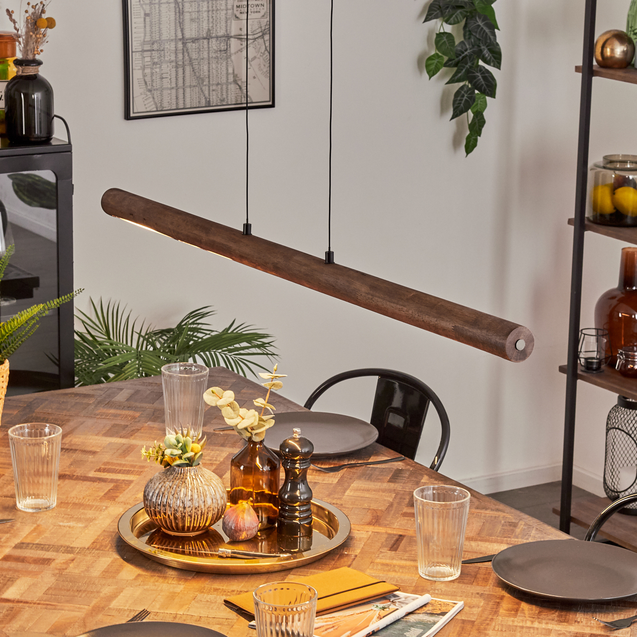 Ein Esszimmer mit Holztisch, Hängelampe und Tablett mit Deko. Teller, Gläser und eine Karte an der Wand sind sichtbar.