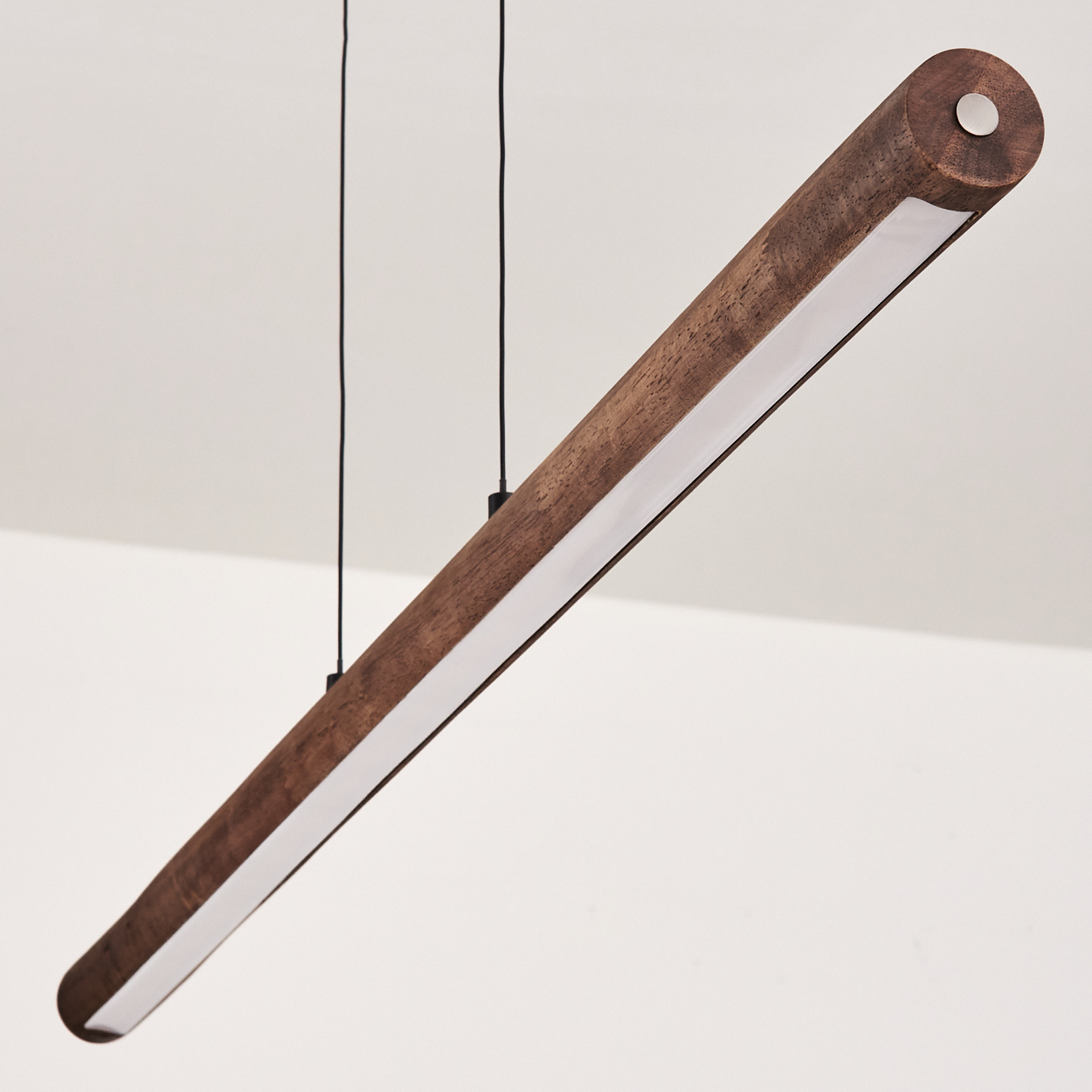Eine zylindrische Holzleuchte hängt an schwarzen Kabeln an einer weißen Decke. Das Licht hat eine helle, weiße Platte.
