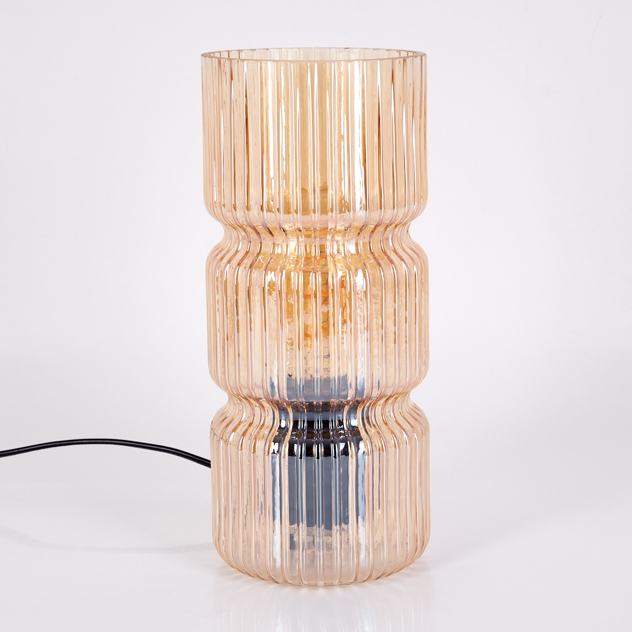 Eine beige, gerippte Glaslampe auf einer weißen Oberfläche, schwarzes Kabel. Die Lampe hat eine zylindrische Form mit drei gestapelten Abschnitten.