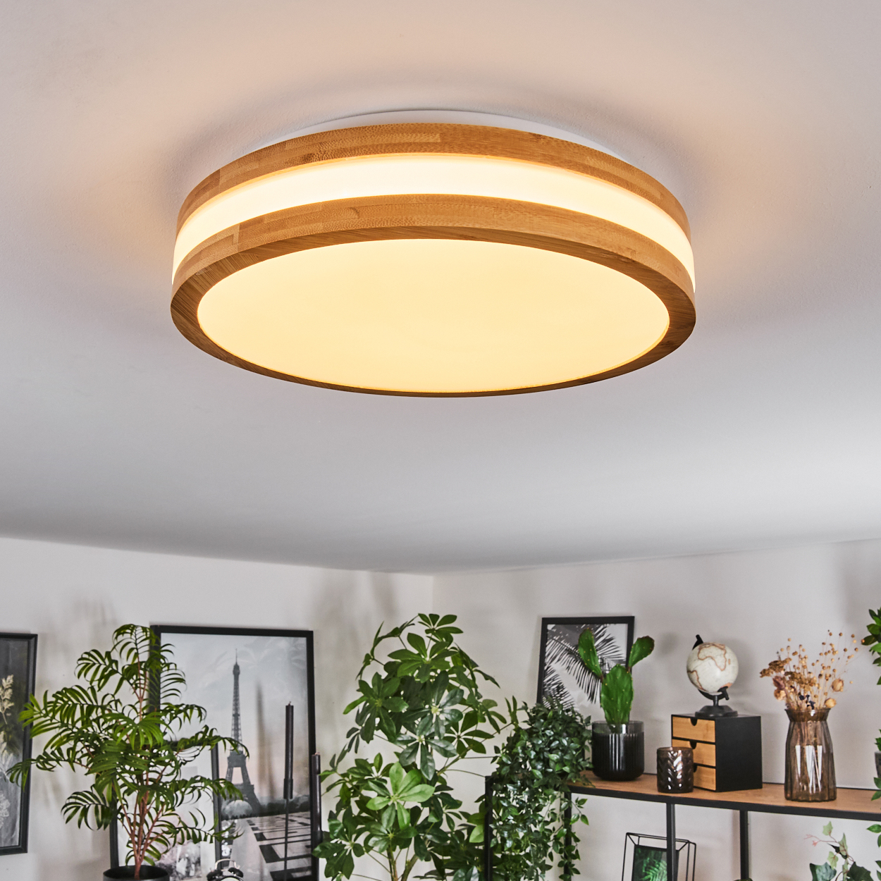 Eine moderne Deckenlampe mit Holzrahmen und hellem Licht. Der Hintergrund zeigt Pflanzen, Bilder und ein Regal.