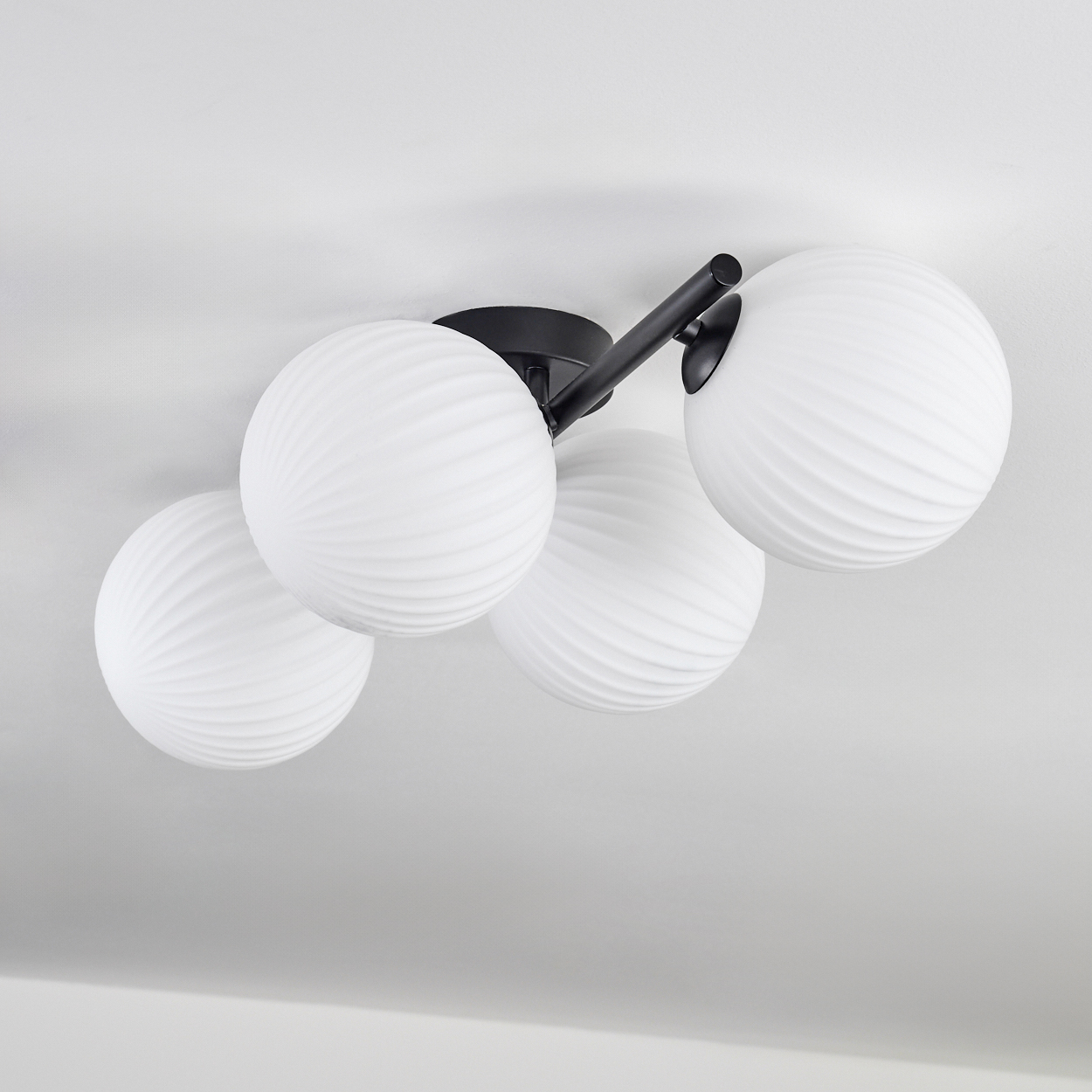 Eine Deckenleuchte mit vier weißen, kugelförmigen Lampen. Die Leuchte hat einen schwarzen Metallrahmen und ist an einer weißen Decke montiert.