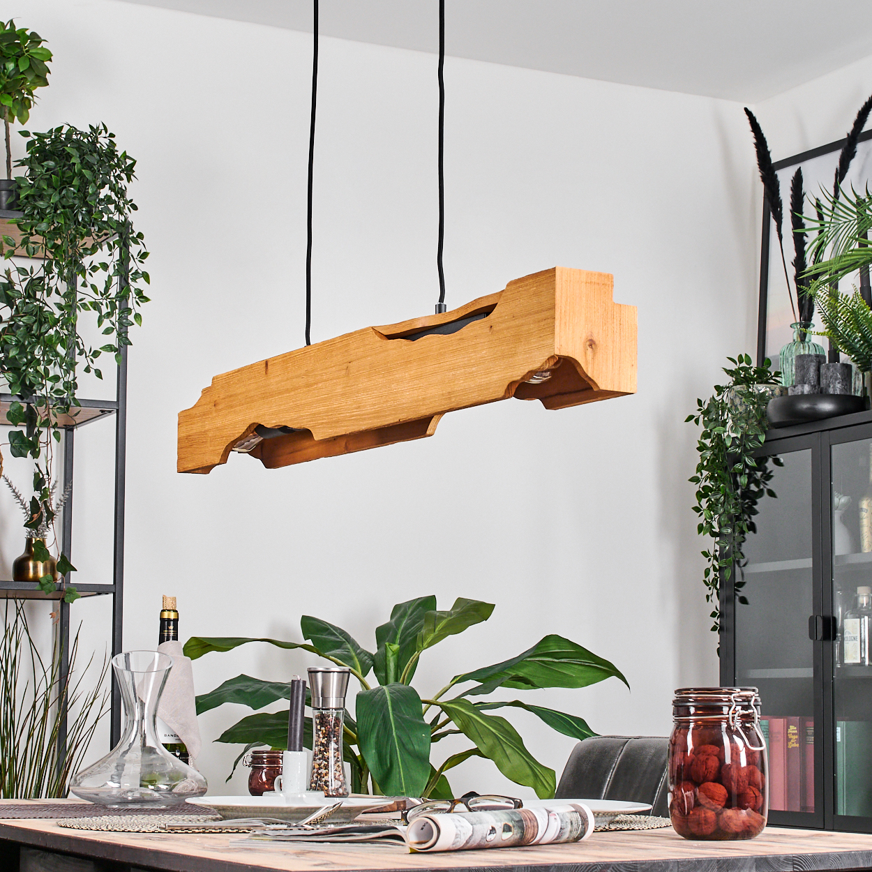 Holzbalken-Deckenleuchte über einem Esstisch. Pflanzen, Glas mit roten Gegenständen und Geschirr auf dem Tisch.