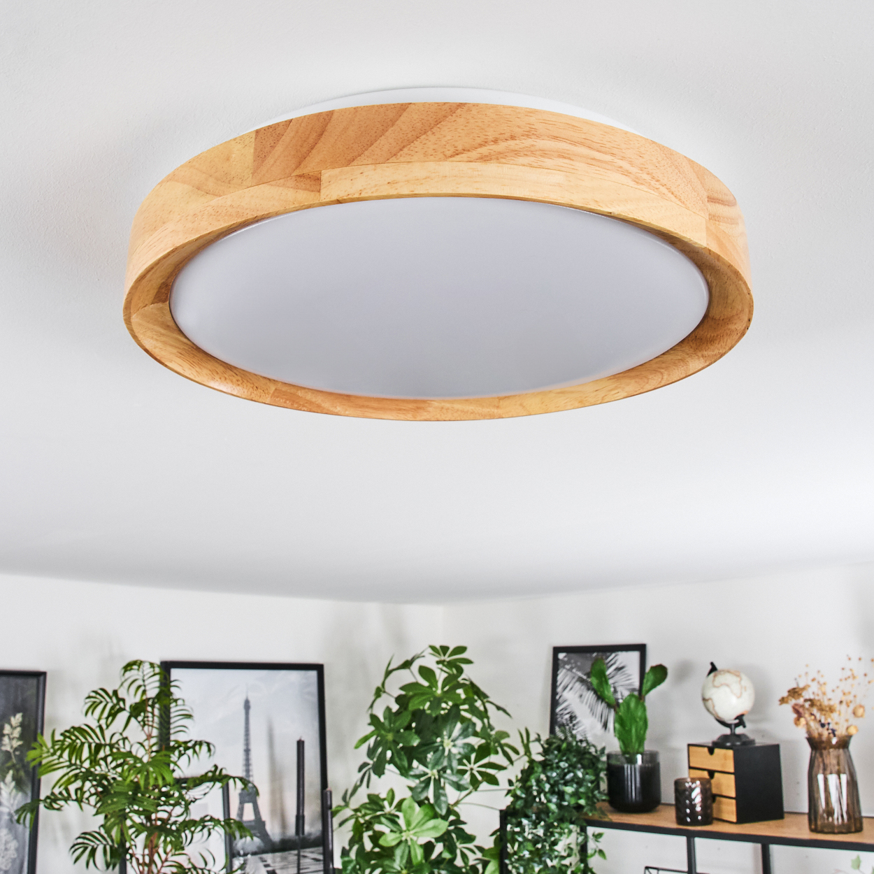 Eine runde Deckenlampe aus Holz mit weißer Mitte. Darunter sind Pflanzen und gerahmte Bilder zu sehen.