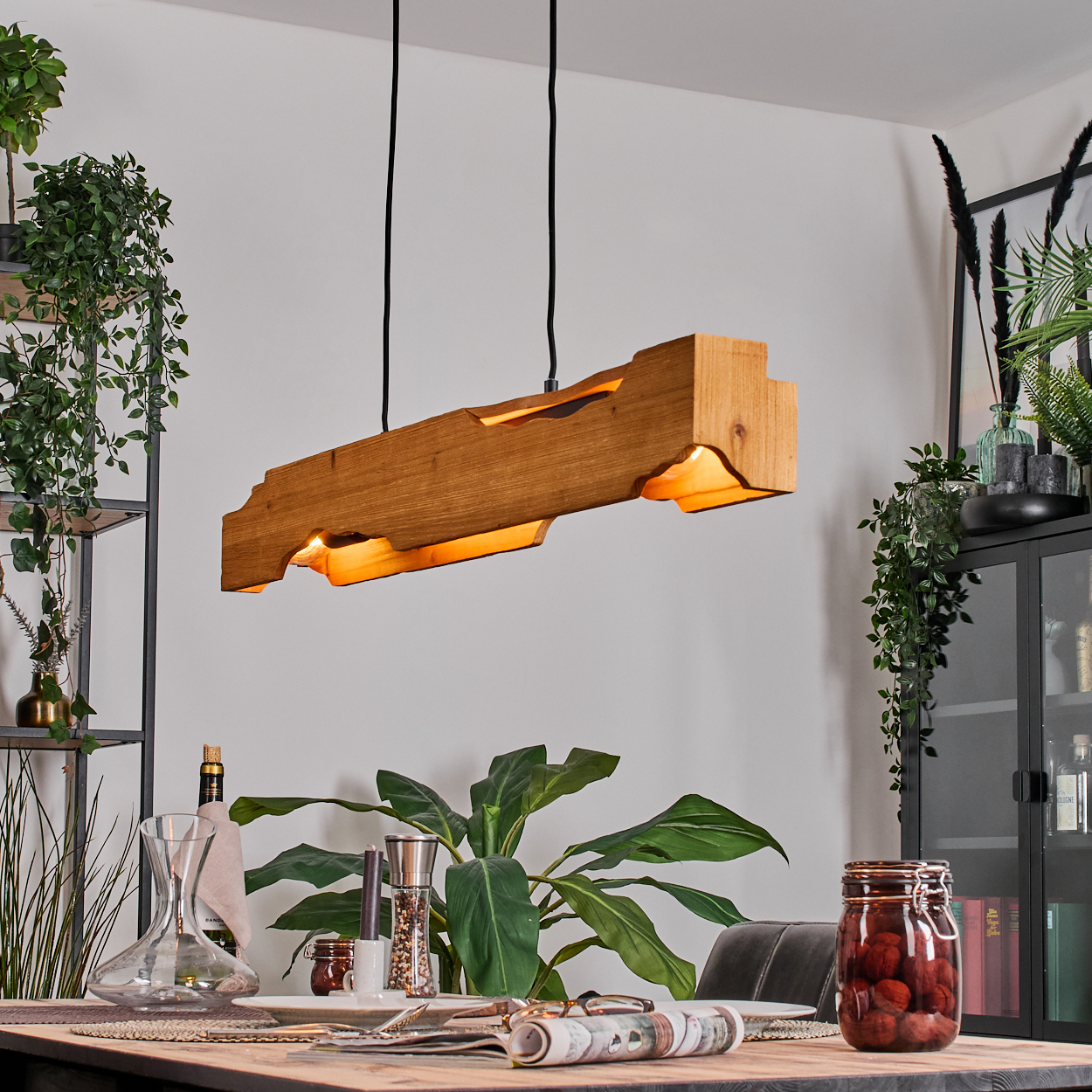Eine Holz-Deckenleuchte hängt über einem Esstisch. Pflanzen umgeben den Tisch, und ein Glas mit roten Kugeln steht darauf.
