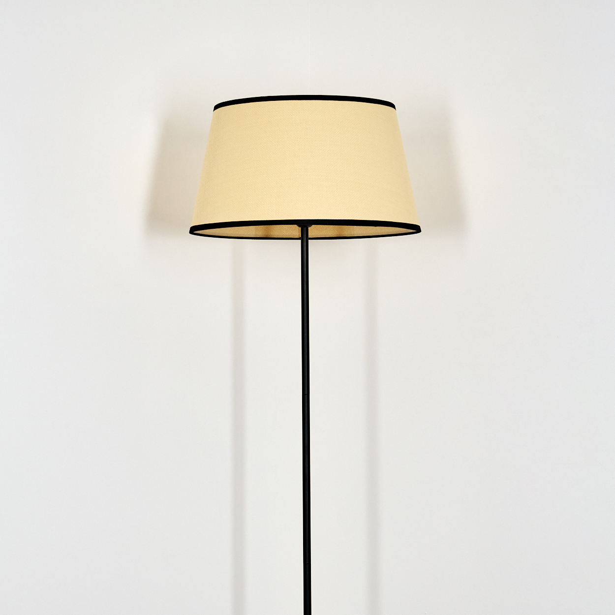 Eine Stehlampe mit beigem Lampenschirm und schwarzer Umrandung steht vor einer weißen Wand. Die Lampe hat einen schwarzen Fuß und Stiel.