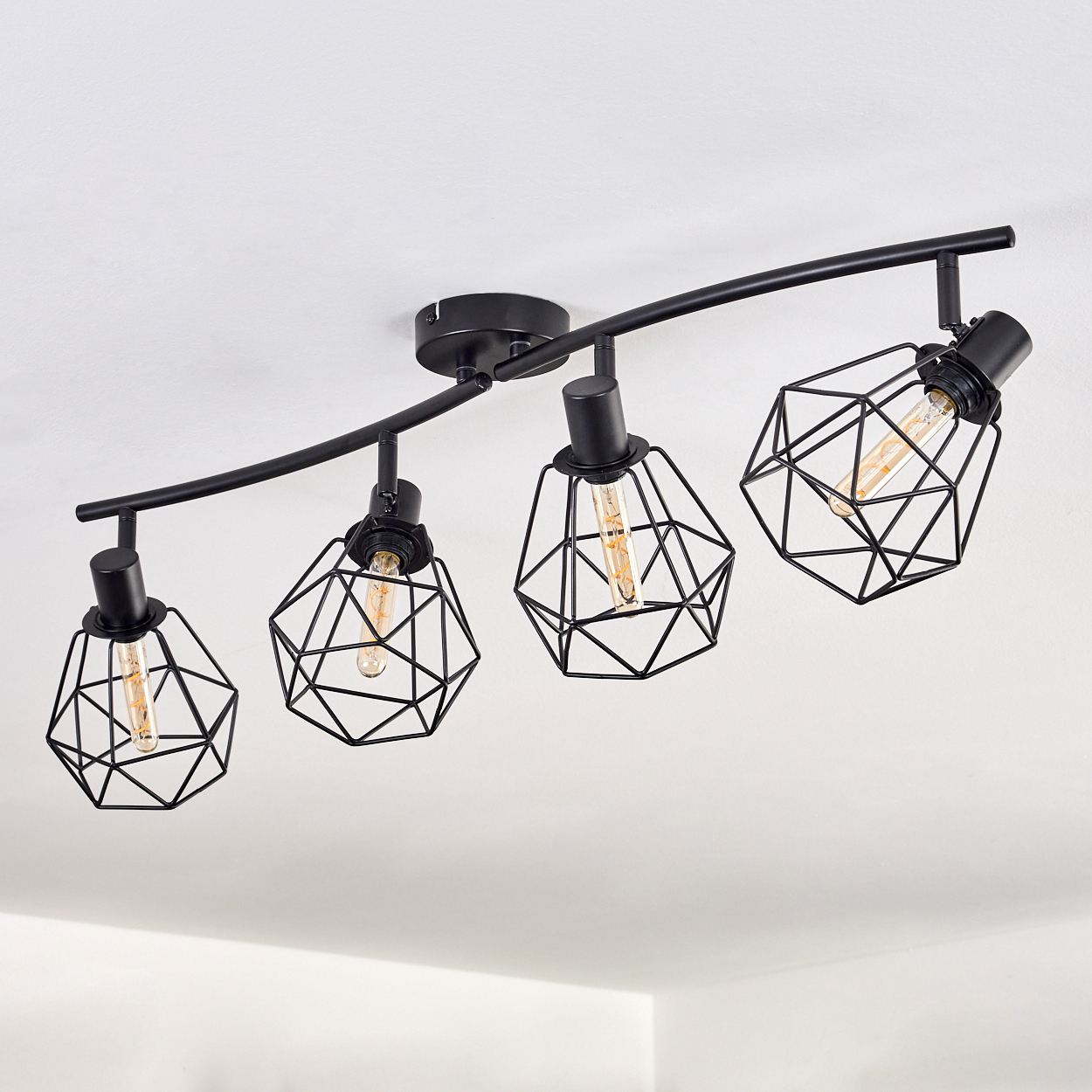 Vier schwarze geometrische Lampen hängen von einer weißen Decke. Jede hat eine Glühbirne.