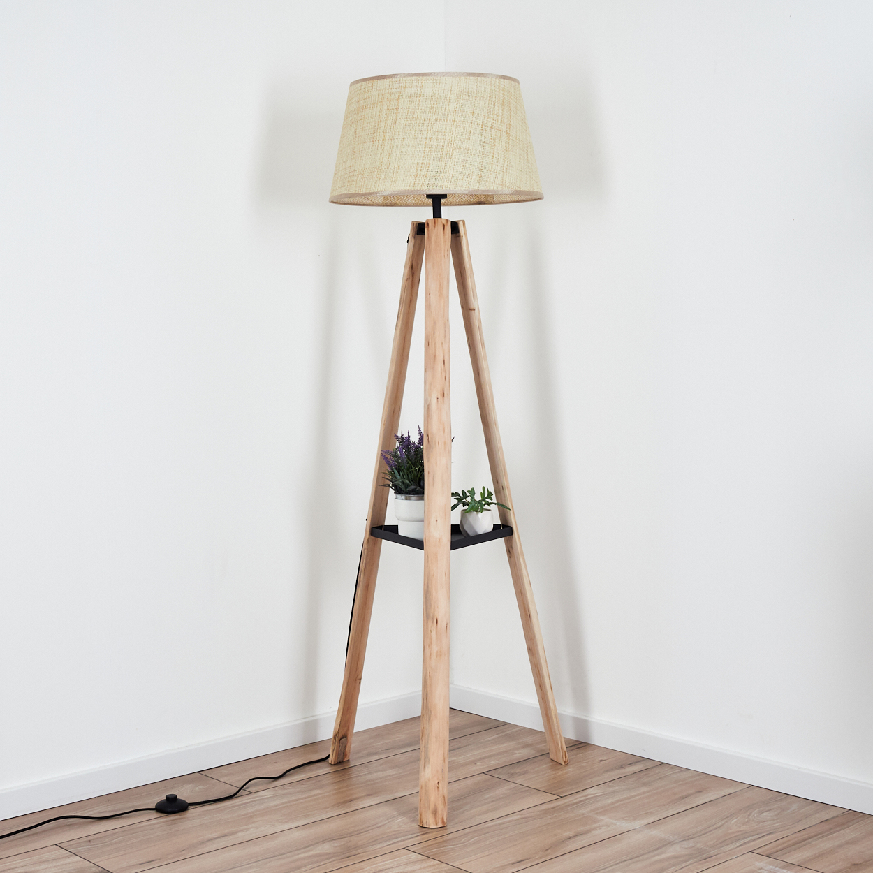 Eine hölzerne Stativ-Stehlampe mit beigem Lampenschirm. Sie hat ein schwarzes Regal mit zwei kleinen Topfpflanzen. Der Boden ist aus Holz.