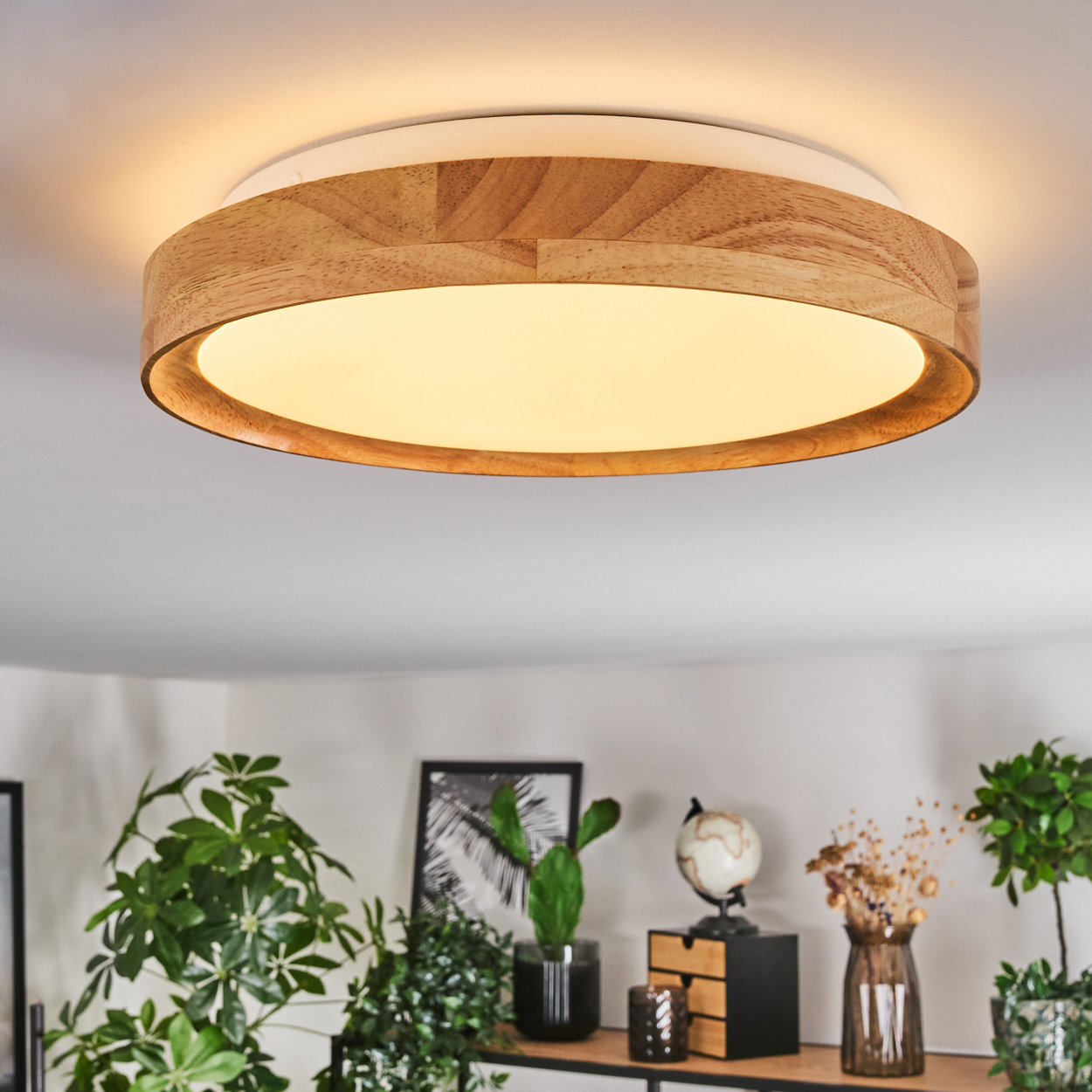 Eine runde Deckenlampe aus Holz beleuchtet einen Raum. Darunter stehen Pflanzen, ein Globus und Dekorationsartikel auf einem Regal.