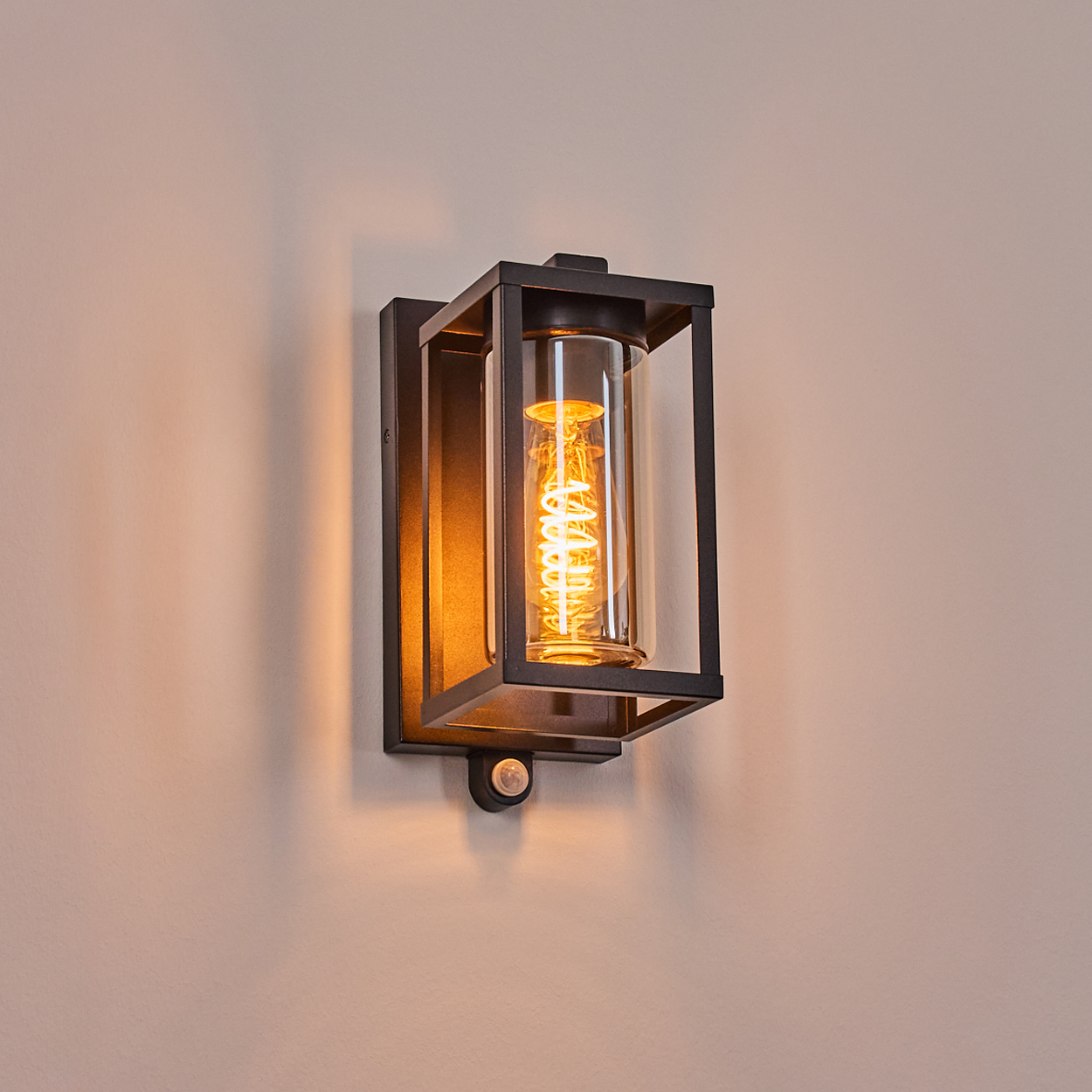 Schwarze Metall-Wandlampe mit einem Glaszylinder, der eine gewendelte Glühbirne enthält. Die Lampe ist an einer beigefarbenen Wand befestigt.