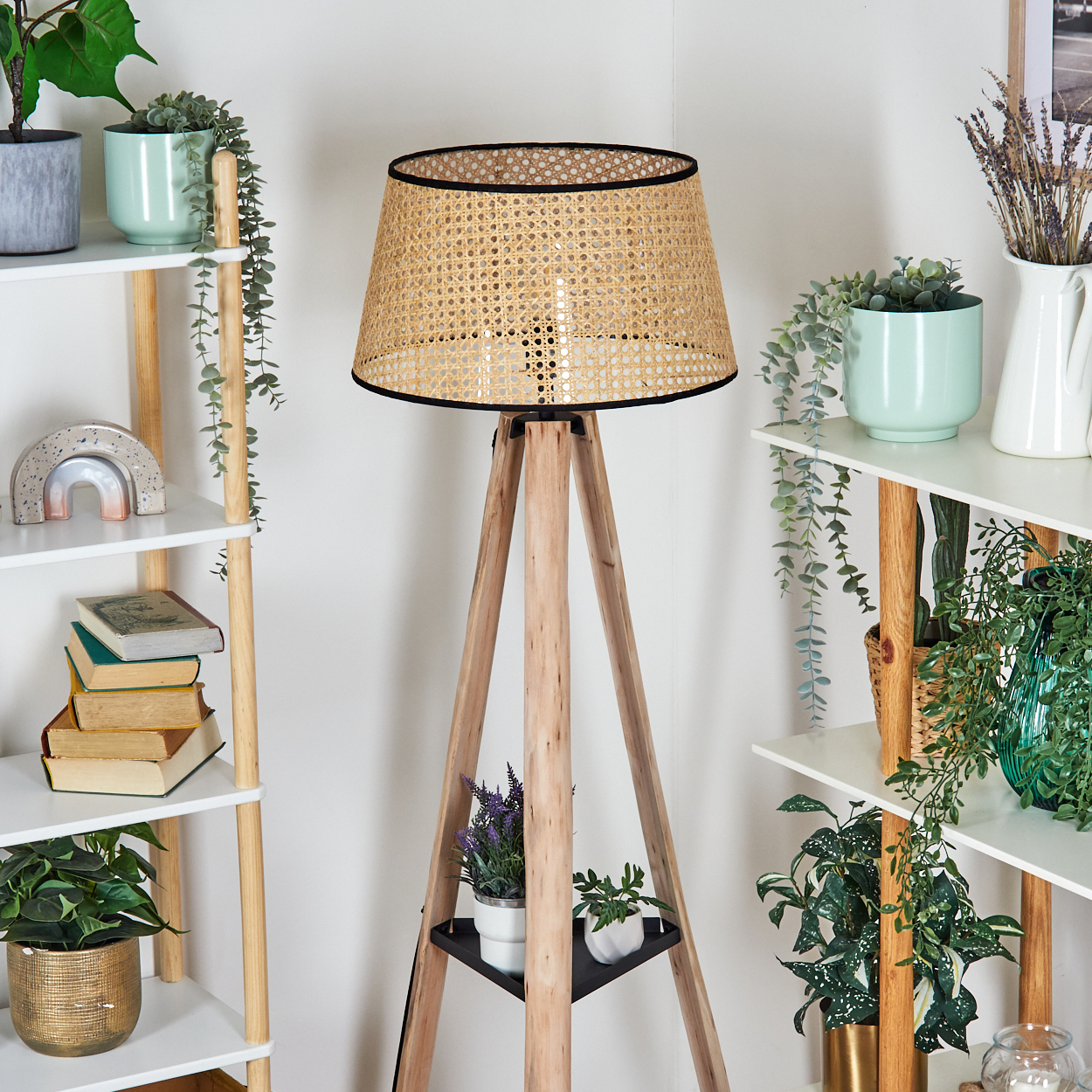 Eine hölzerne Stehlampen-Stativlampe mit einem gewebten Schirm steht vor weißen Regalen mit Pflanzen, Büchern und Dekorationsartikeln.