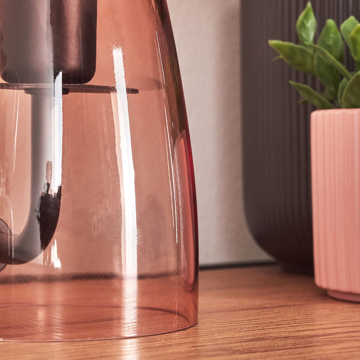 Ein rosa Glasobjekt mit dunkler Basis steht auf einer Holzoberfläche. Rechts ist eine grüne Pflanze in einem rosa Topf.
