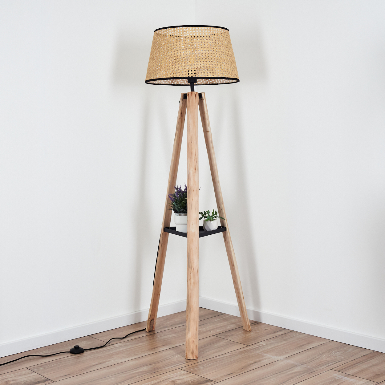 Eine hölzerne Stativ-Stehlampe mit gewebtem Lampenschirm. Ein Regal hält zwei kleine Topfpflanzen. Der Boden ist aus Holz und die Wand ist weiß.