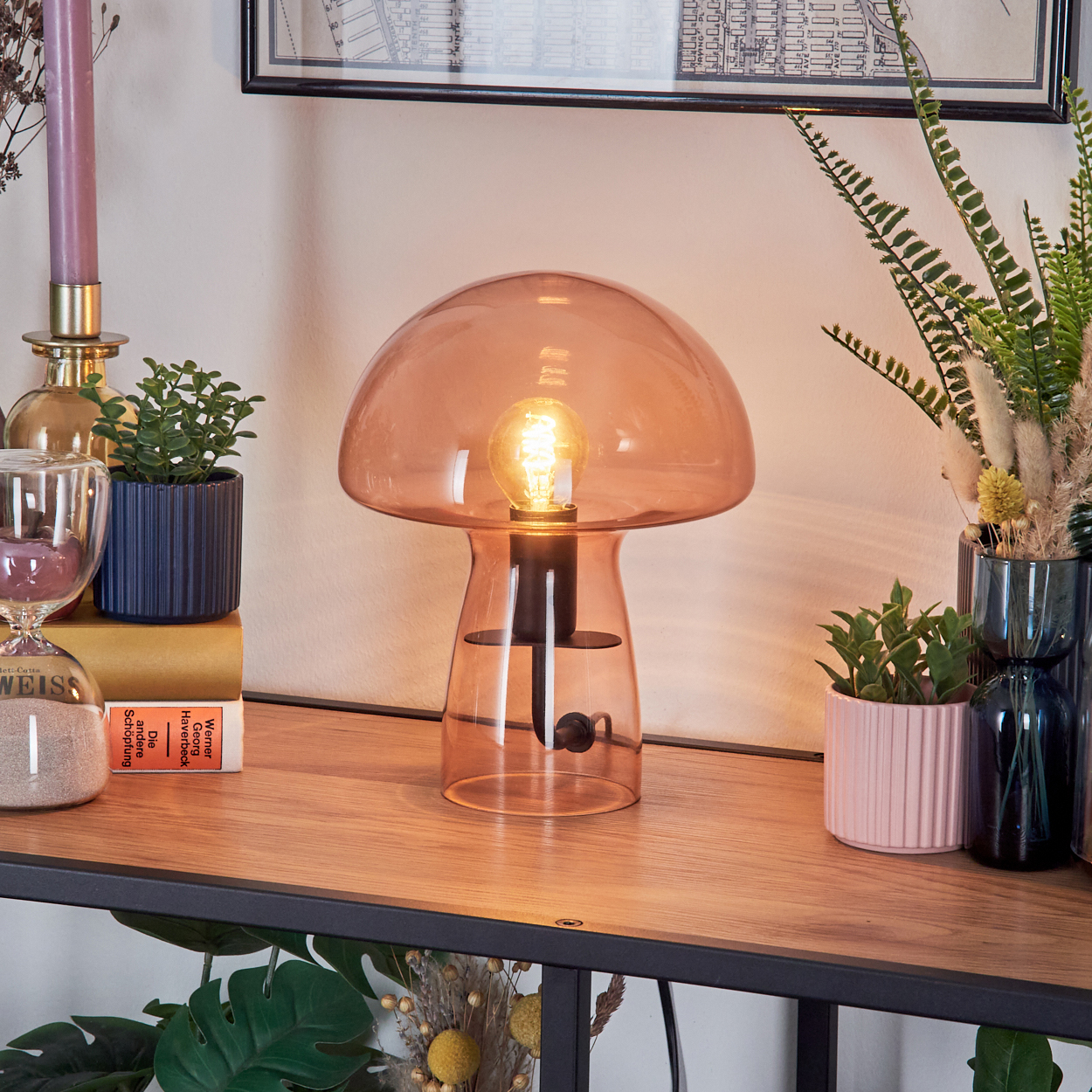 Eine pilzförmige Lampe leuchtet auf einem Holzregal mit Pflanzen, Büchern und einer gerahmten Karte im Hintergrund. Sanduhr und Kerze sind ebenfalls vorhanden.