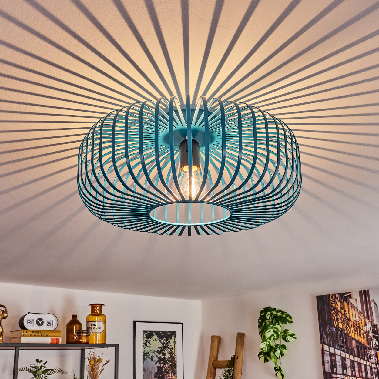 Eine türkisfarbene, käfigartige Deckenlampe wirft Schatten auf die weiße Decke. Darunter sind ein dekoriertes Regal und gerahmte Kunst zu sehen.