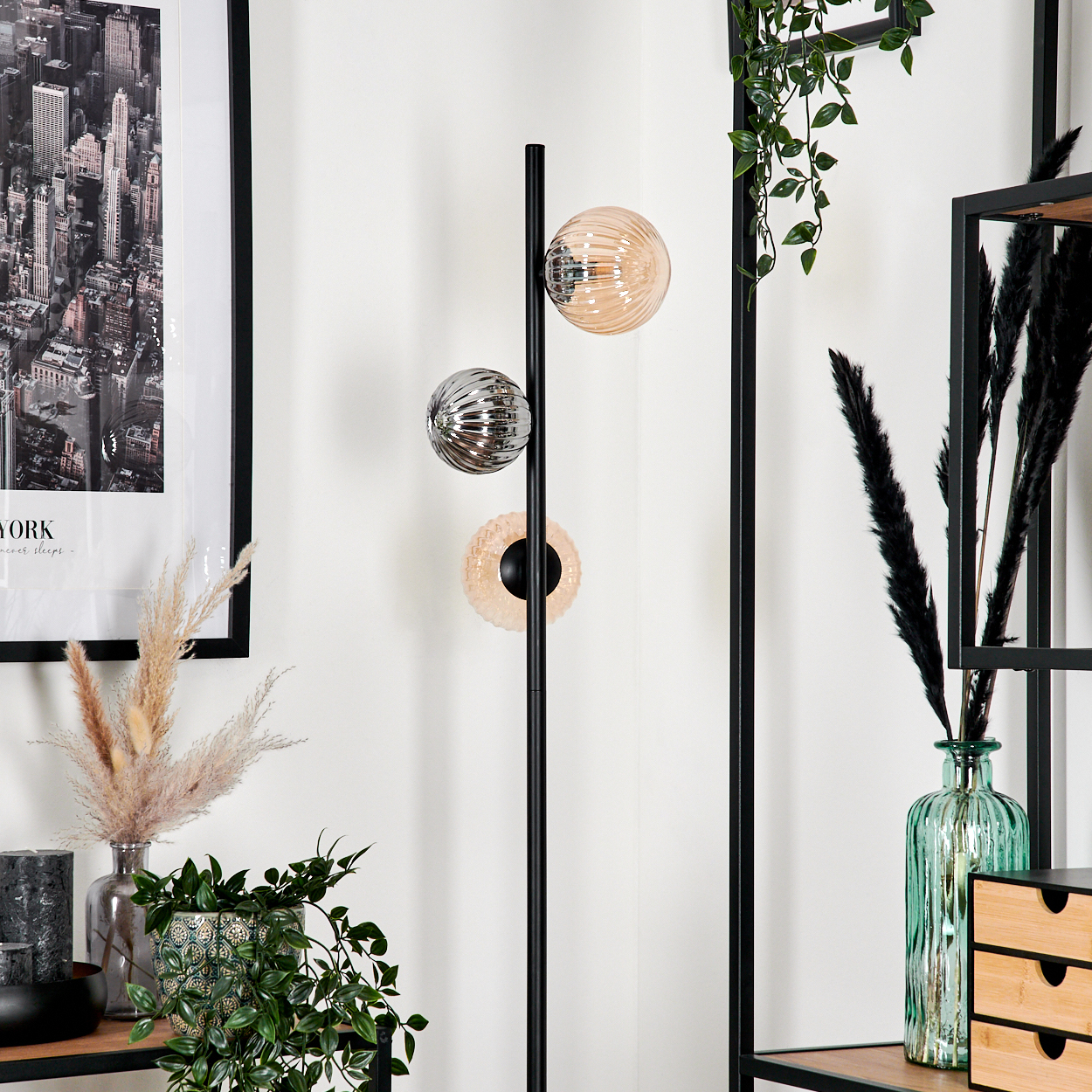 Ein modernes Interieur mit einer schwarzen Stehlampe mit drei kugelförmigen Lichtern. Auf den Regalen und an der Wand befinden sich Pflanzen und Dekorationen.