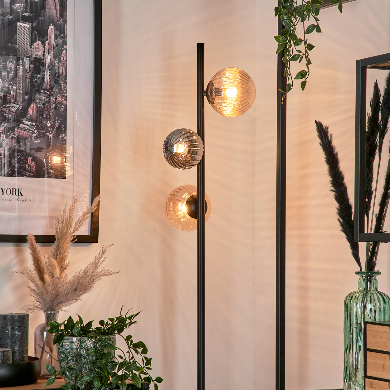 Eine moderne Stehlampe mit drei Glaskugelleuchten. Im Hintergrund ein gerahmtes Stadtbild und dekorative Pflanzen.