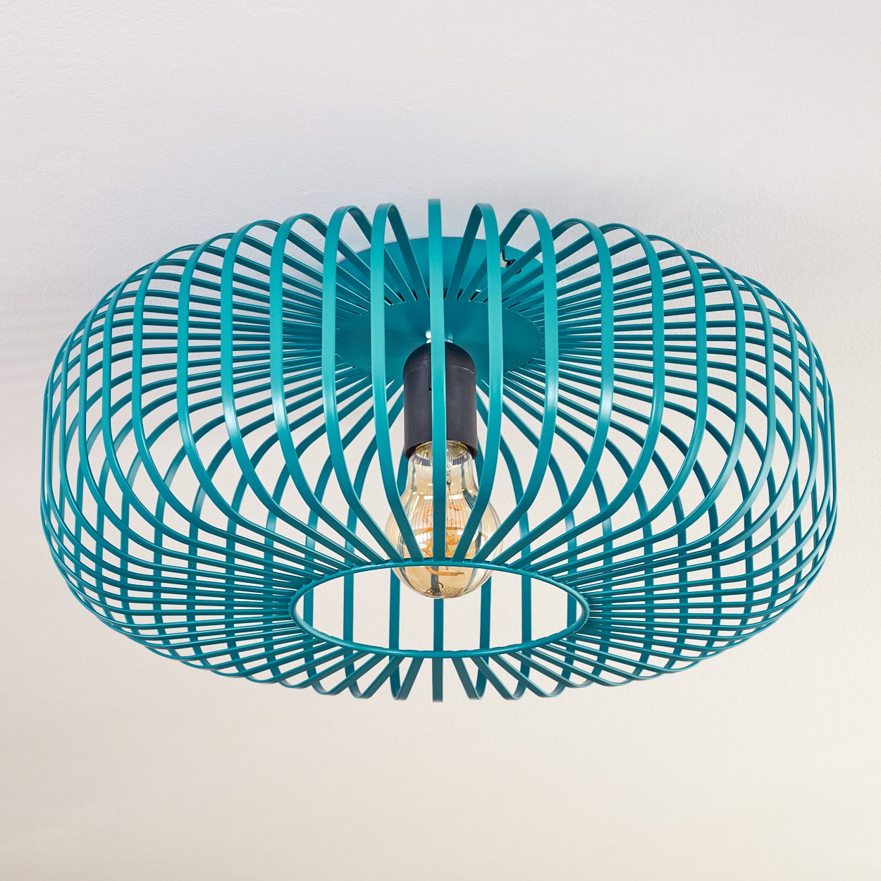 Eine teal-farbene, käfigartige Deckenlampe mit sichtbarer Glühbirne. Der Hintergrund ist eine schlichte weiße Decke.