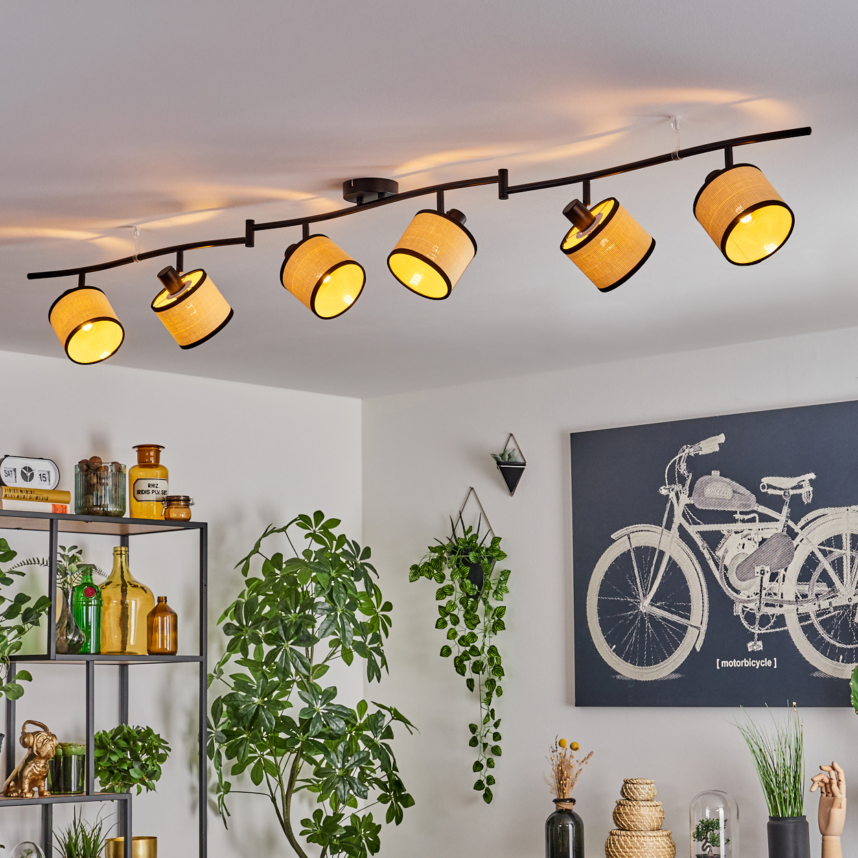 Eine Deckenlampe mit mehreren Lampenschirmen ist beleuchtet. Der Raum hat Regale mit Dekor, Pflanzen und eine Fahrradzeichnung an der Wand.