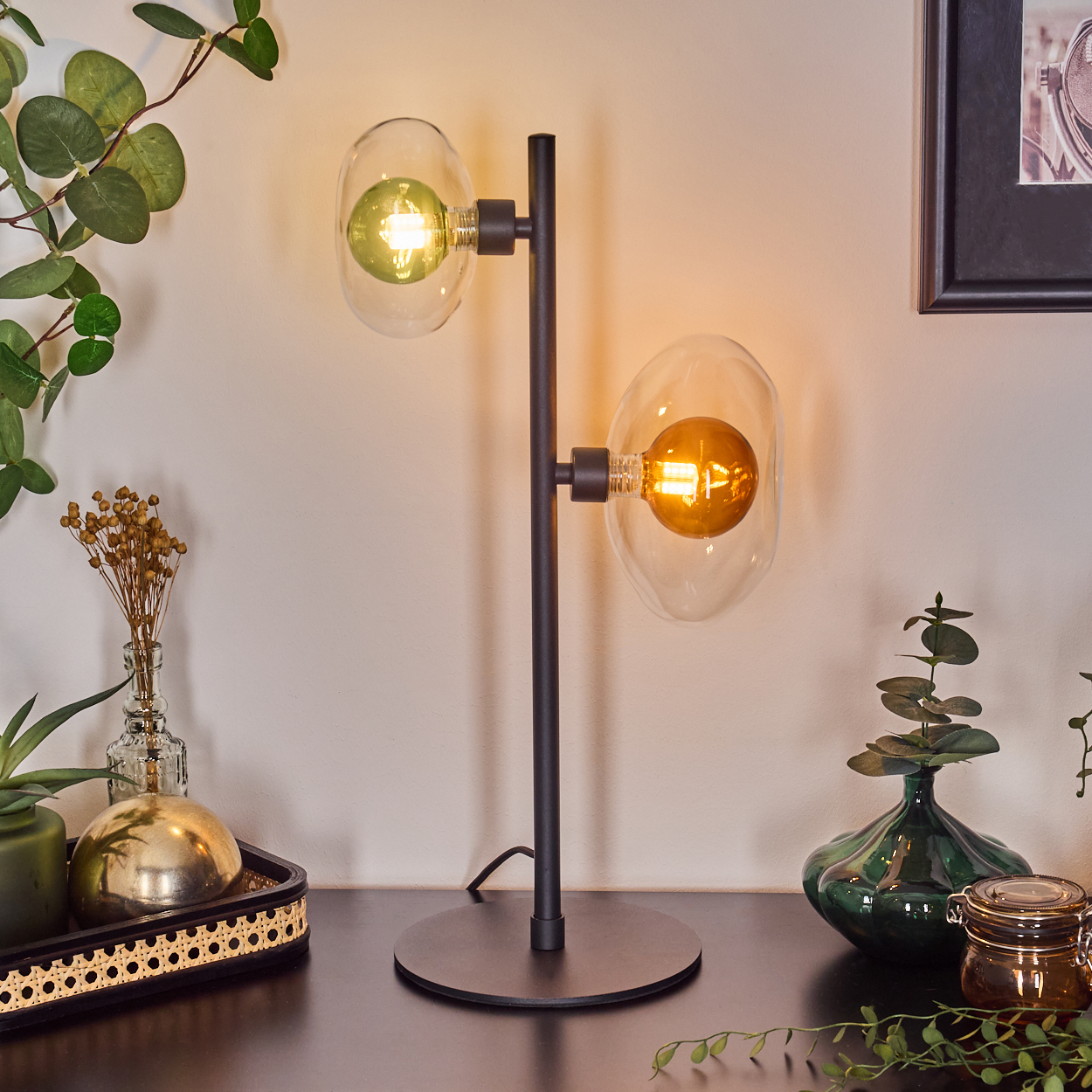 Eine schwarze Lampe mit zwei Glaskolbenhaltern, einer grünen und einer orangen Glühbirne, steht auf einem Tisch mit Pflanzen und einem gerahmten Bild.