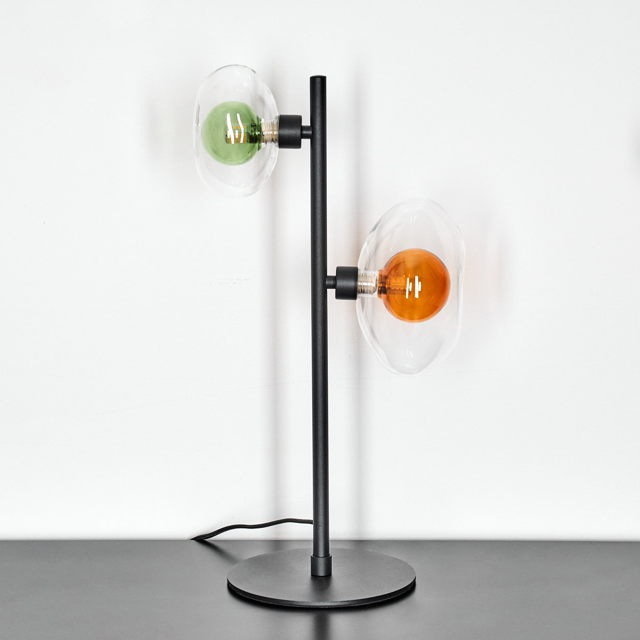 Eine moderne Lampe mit schwarzem Ständer und zwei Glasschirmen. Eine hat eine grüne Glühbirne, die andere eine orangefarbene. Der Hintergrund ist weiß.