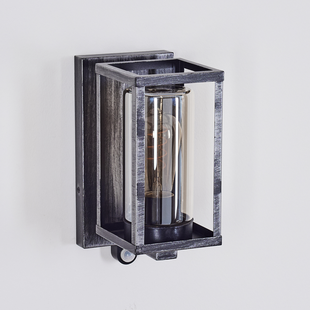 Eine Wandleuchte für den Außenbereich. Sie hat einen rechteckigen Rahmen um einen Glaszylinder, in dem sich eine Glühbirne befindet. Die Wand ist weiß.