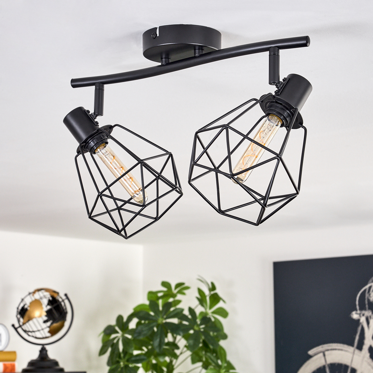 Eine schwarze Deckenlampe mit zwei Lichtern. Jedes Licht befindet sich in einem geometrischen Käfig. Ein Globus und eine Pflanze sind im Hintergrund.