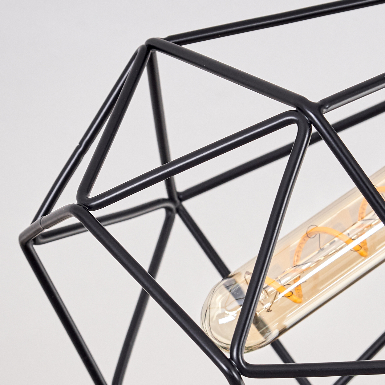 Nahaufnahme einer schwarzen, geometrischen, käfigartigen Lampe mit einer langen, bernsteinfarbenen Glühbirne. Der Hintergrund ist weiß.