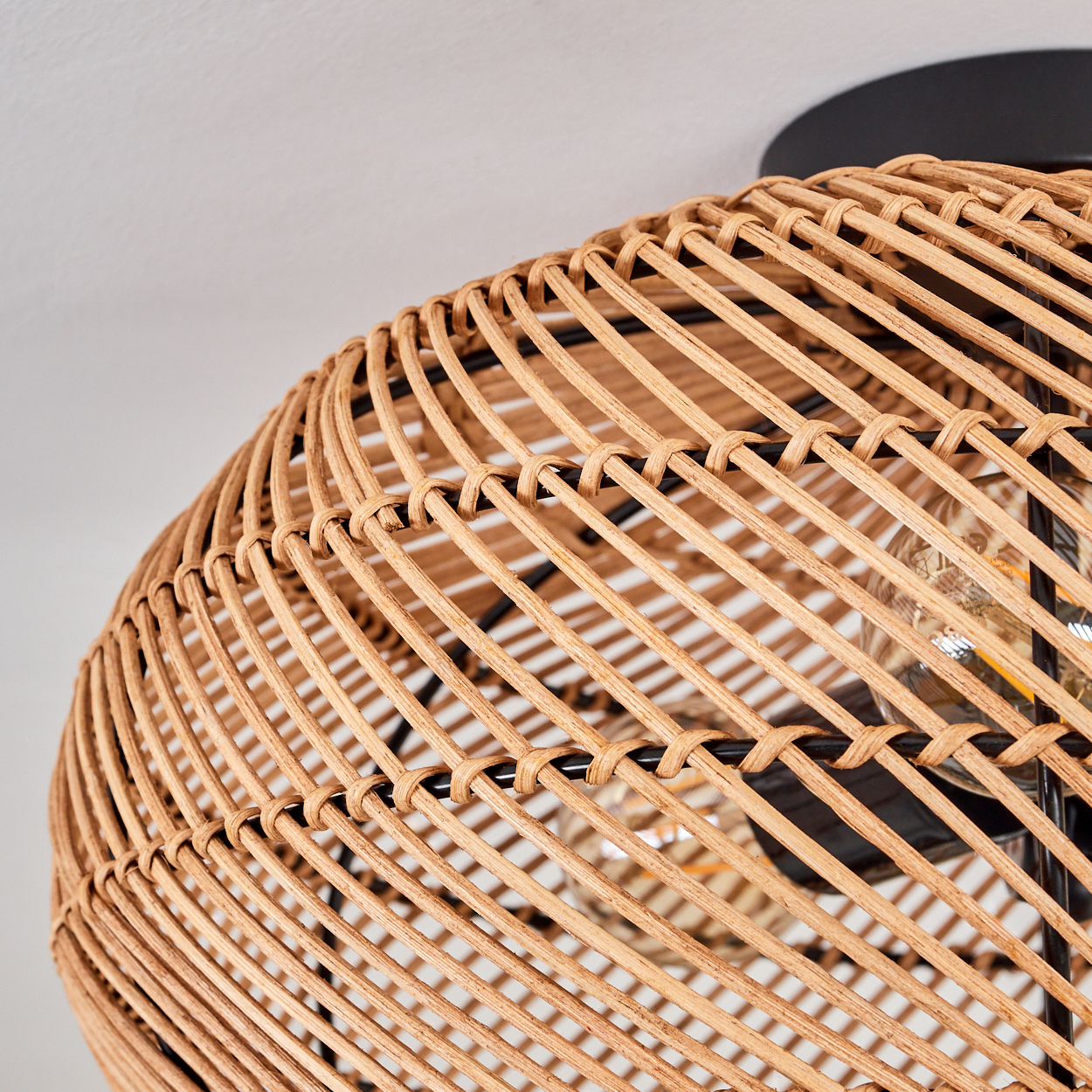 Nahaufnahme einer gewebten Pendelleuchte aus Rattan mit schwarzem Metallrahmen. Die Glühbirne ist sichtbar. Die Decke ist weiß.