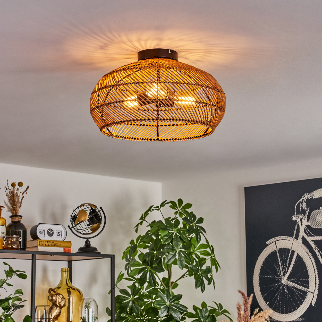 Eine Deckenlampe aus geflochtenem Rattan mit drei Lichtern hängt über einem Raum mit Pflanzen, Büchern und einem Fahrradkunstwerk.