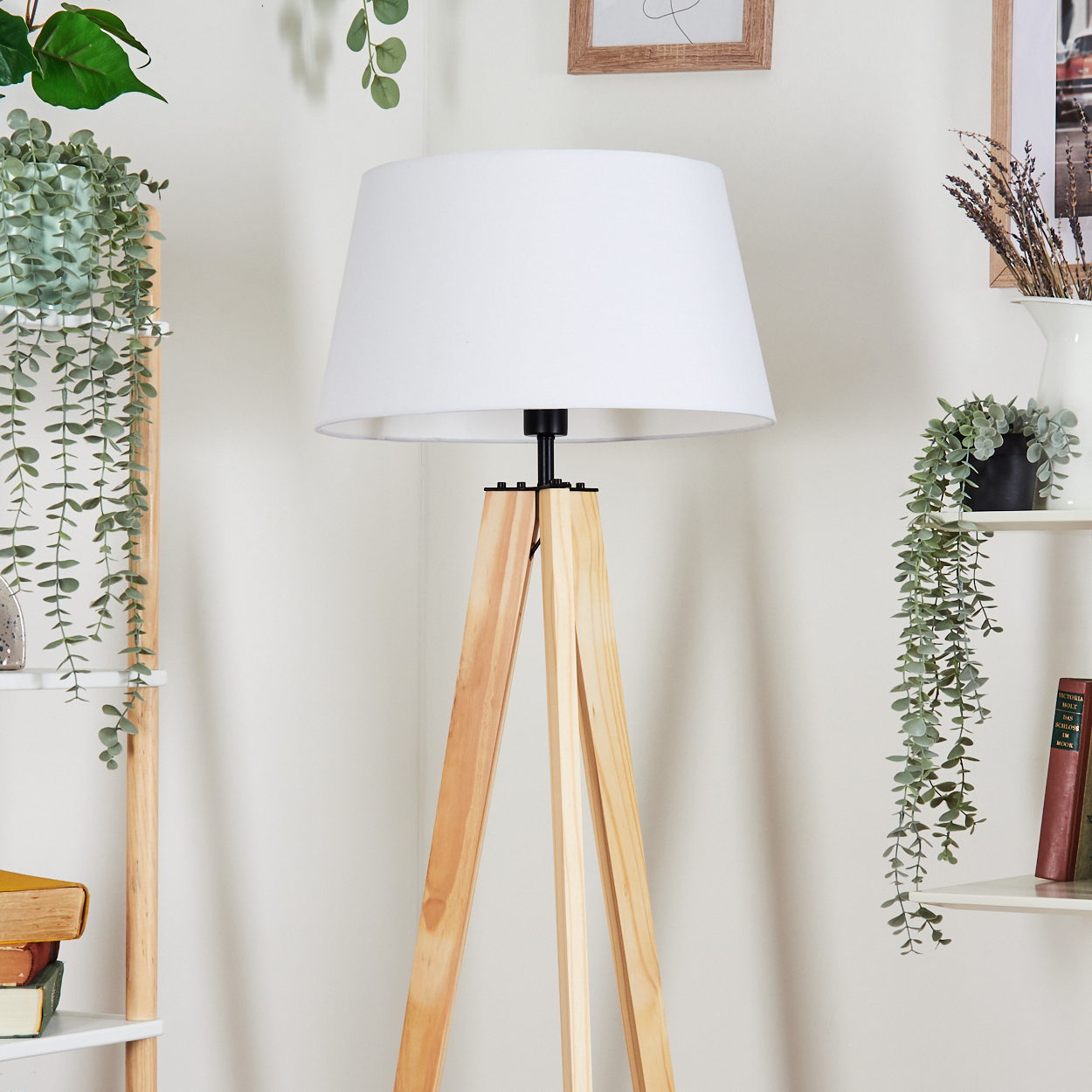 Eine Stehlampe aus Holz mit weißem Schirm, Pflanzen auf Regalen und hängend, sowie gerahmte Bilder im Hintergrund.