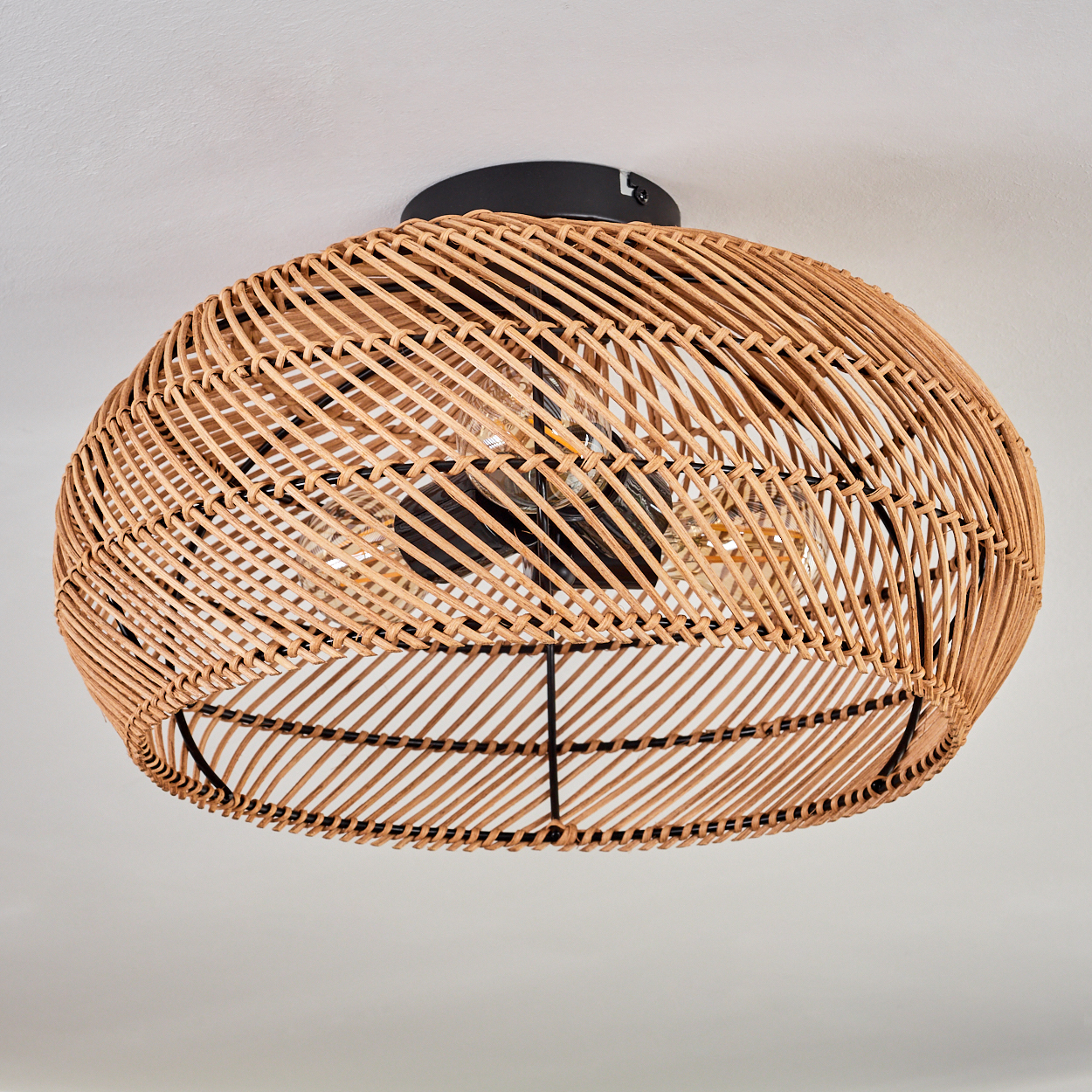 Ein gewebter Lampenschirm aus Rattan hängt von der Decke. Er hat einen schwarzen Sockel und eine Glühbirne ist im Inneren sichtbar.